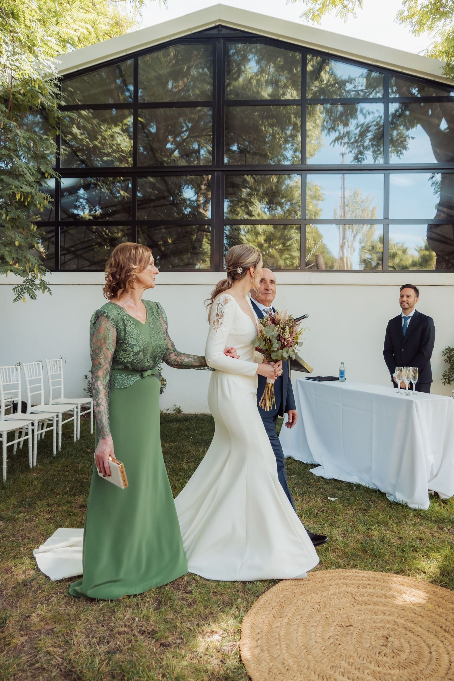 Bride in a fitted white gown walks the aisle escorted by her mother in a green dress.