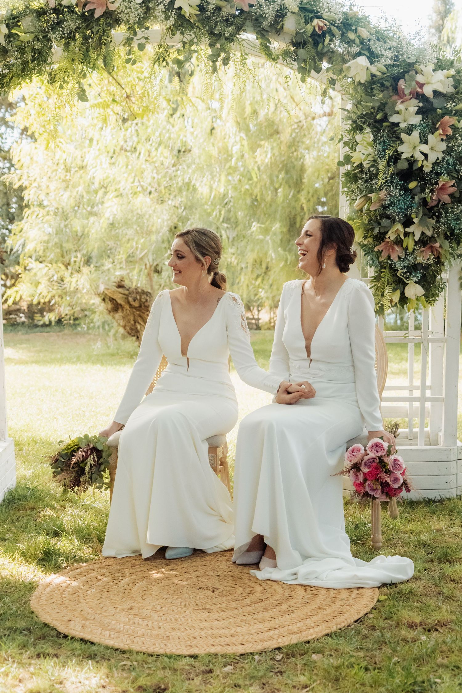 Two brides in white long-sleeve gowns sitting under a floral arch laughing on their wedding day.