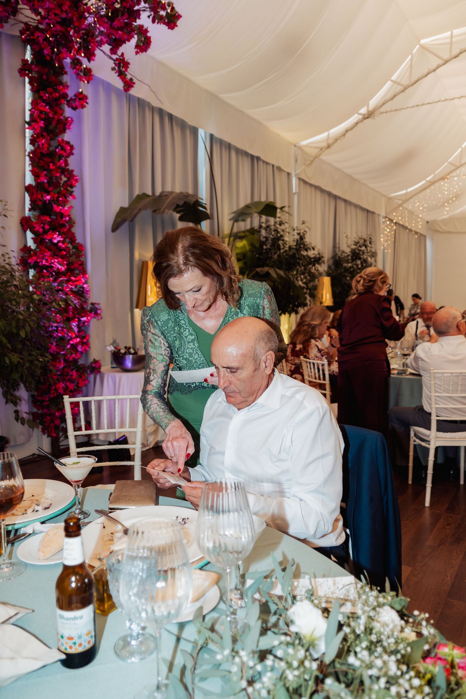 Woman in green lace dress assists seated man at a wedding reception table with floral decorations.