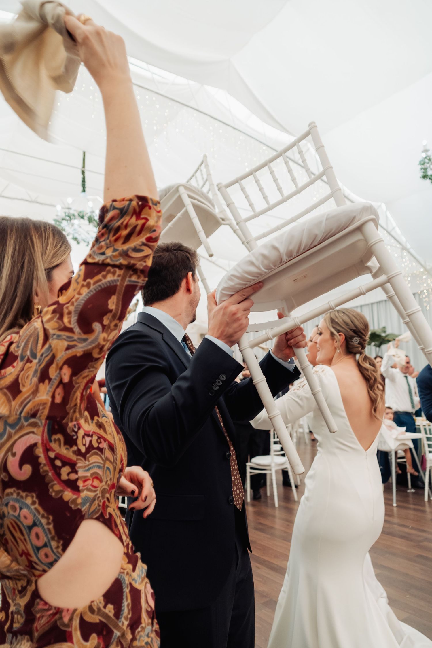 Wedding guests lifting white chairs in celebration as bride in backless gown enters reception tent.
