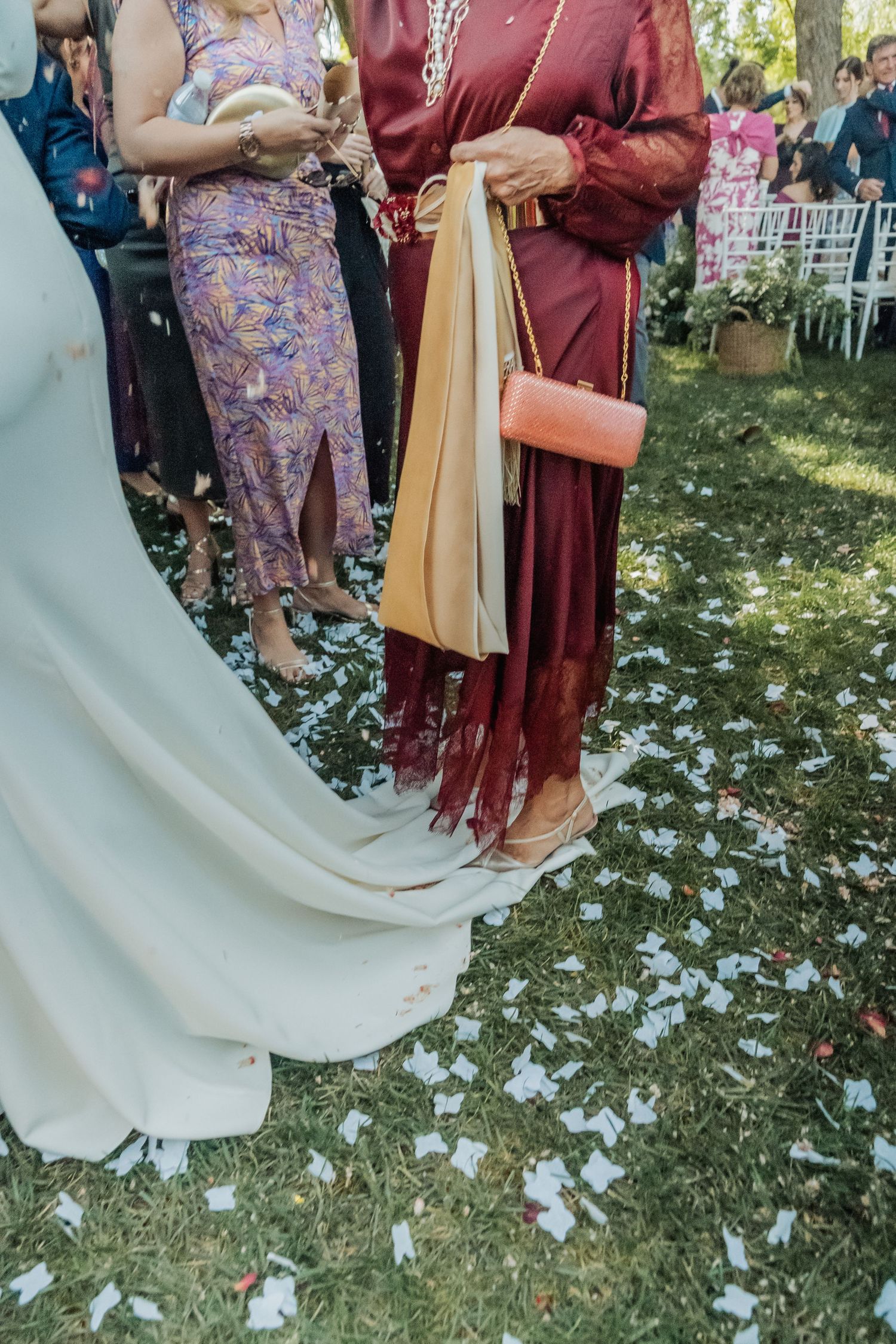 Wedding guests in colorful dresses stand on grass scattered with white confetti petals at outdoor ceremony.