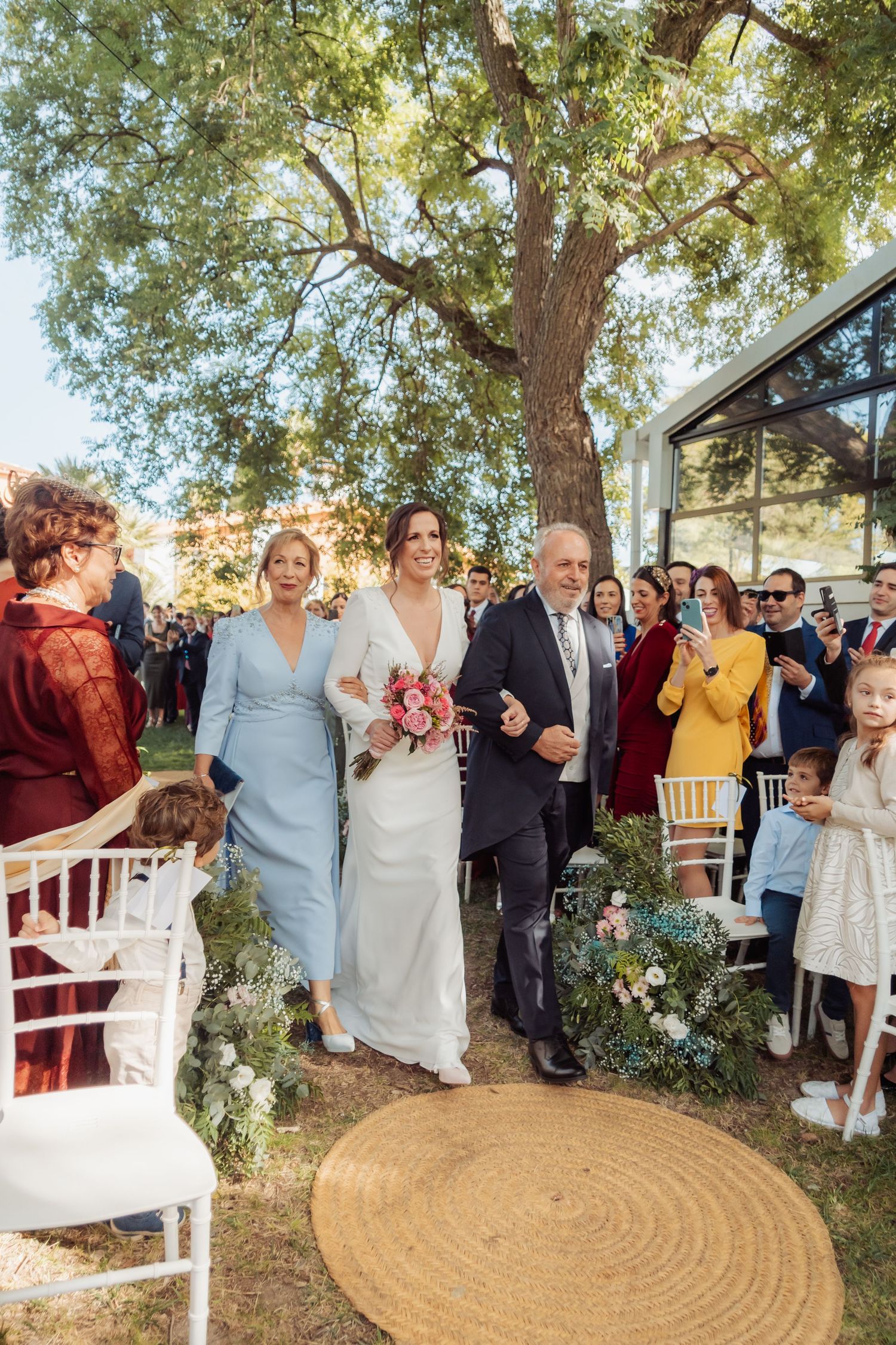 Bride walks down outdoor aisle with parents as wedding guests watch and photograph.