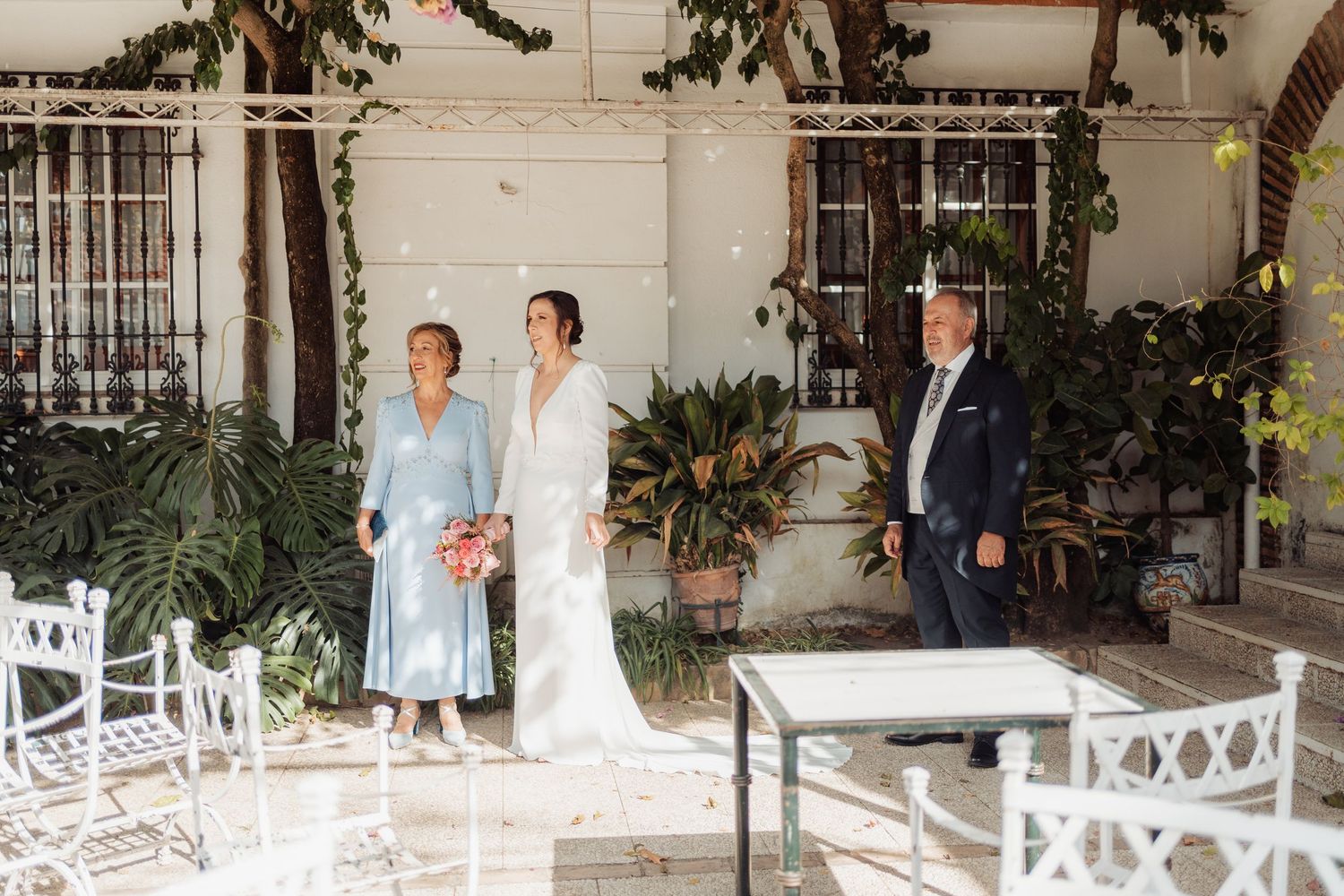 Bride in white gown walks with parents through sunlit courtyard with white garden furniture and green plants.