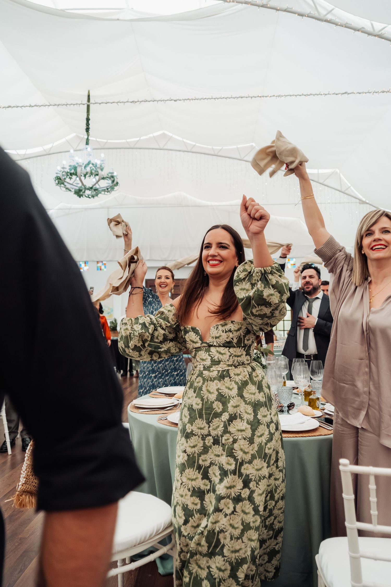Joyful wedding guest in floral green dress celebrating with raised arms inside a white marquee tent.