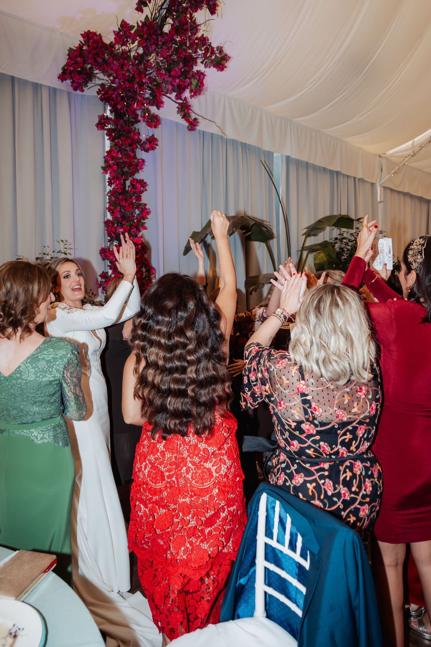 Group of women in colorful dresses celebrating with raised hands at a vibrant wedding reception.