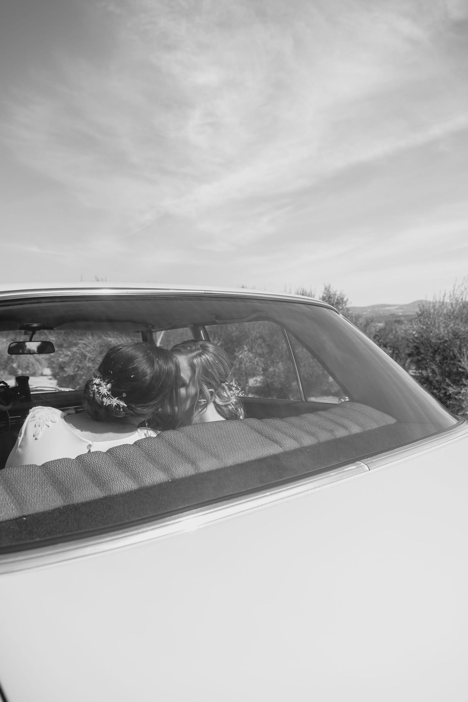 Black and white photo of two brides sharing a kiss inside a vintage white car.