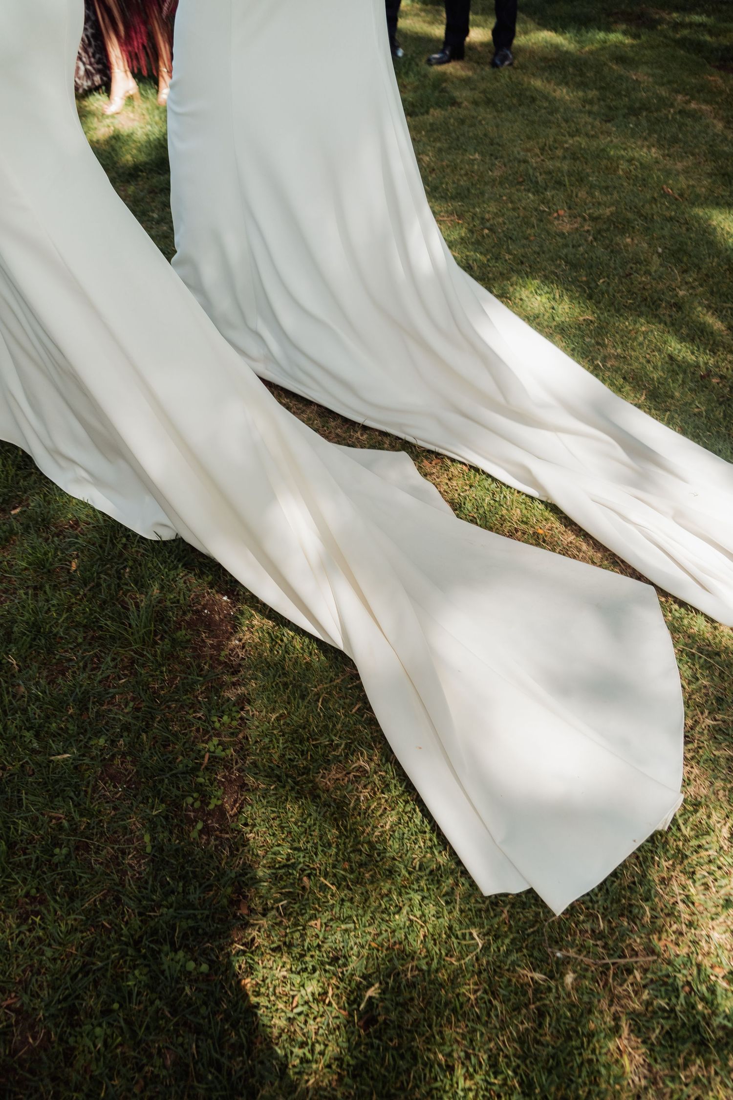 Close-up of a white wedding dress train flowing gracefully over green grass in sunlight.