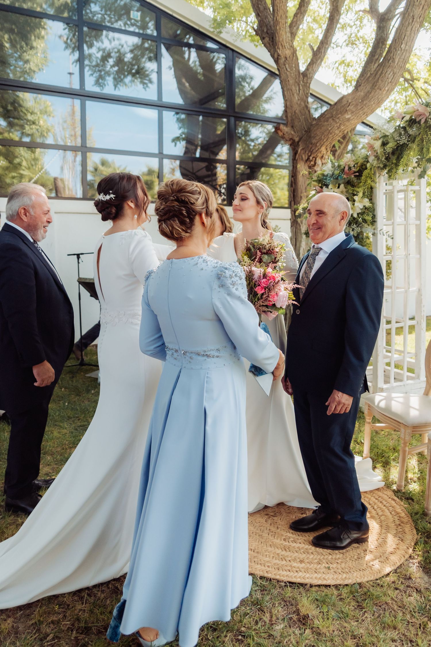 Bride in white gown receives guests at outdoor wedding ceremony with floral arch backdrop.