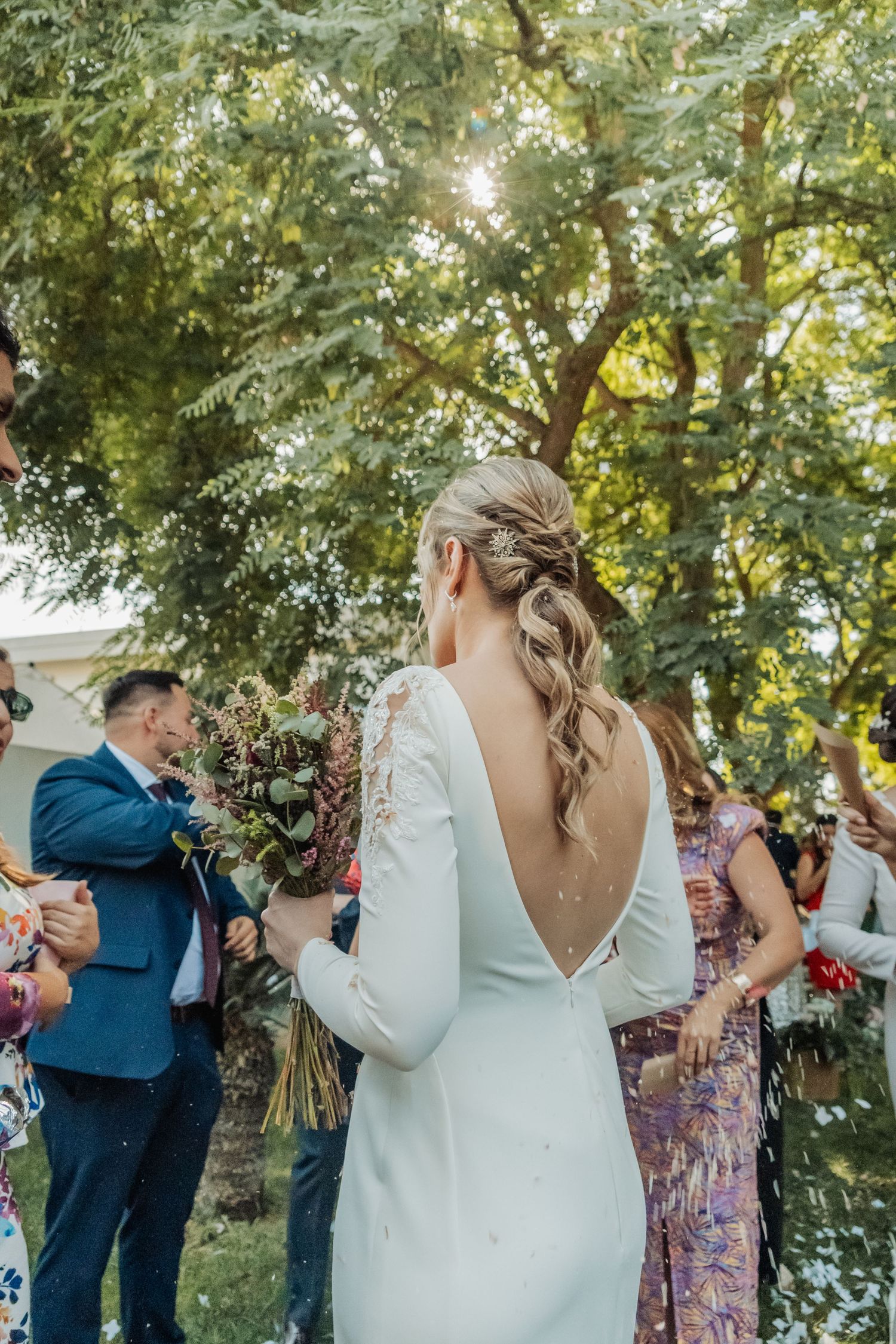 Blonde bride in backless white gown holds bouquet while guests celebrate with confetti at outdoor wedding.