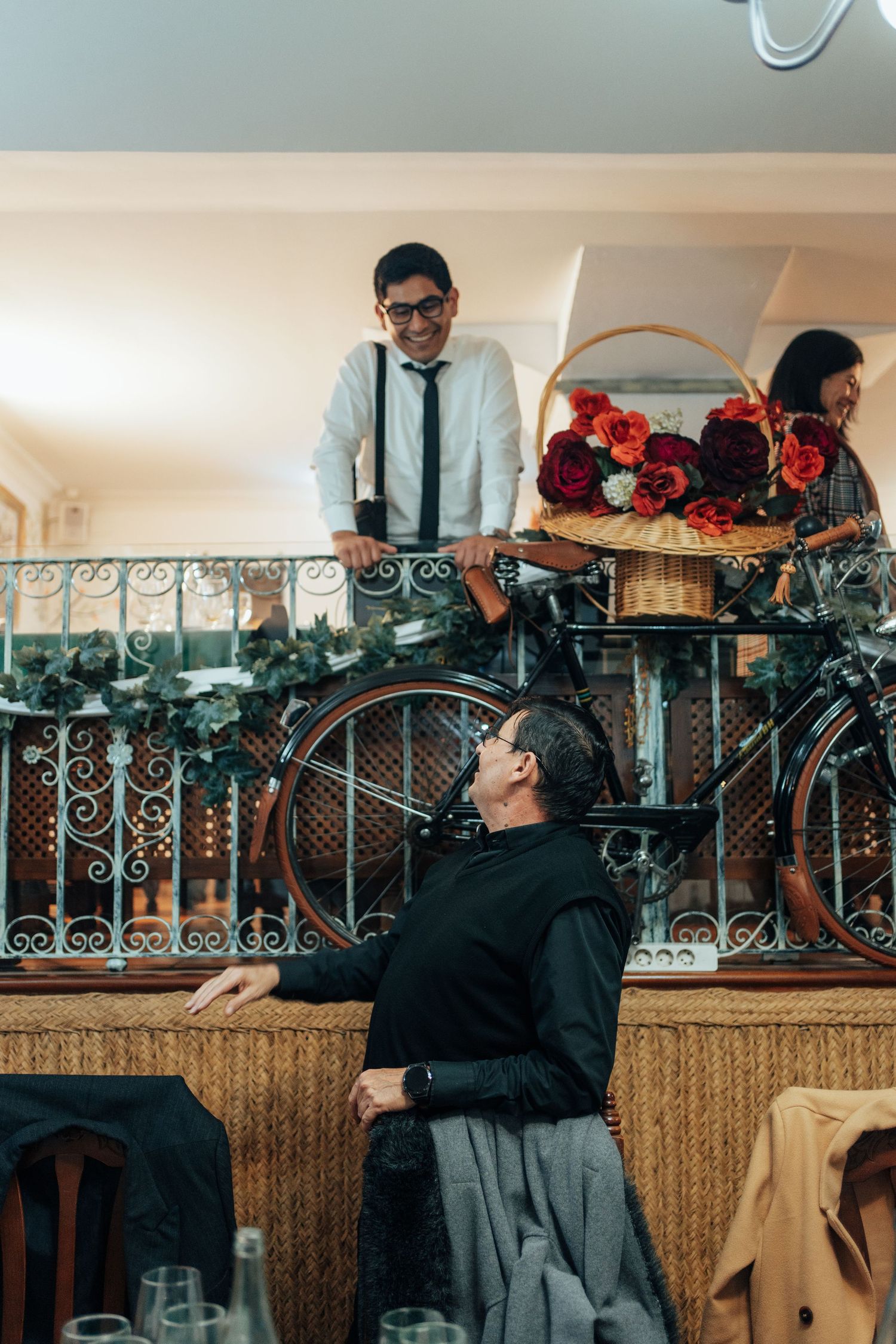 Man in white shirt and tie on balcony with flower basket beside vintage bicycle at wedding venue.