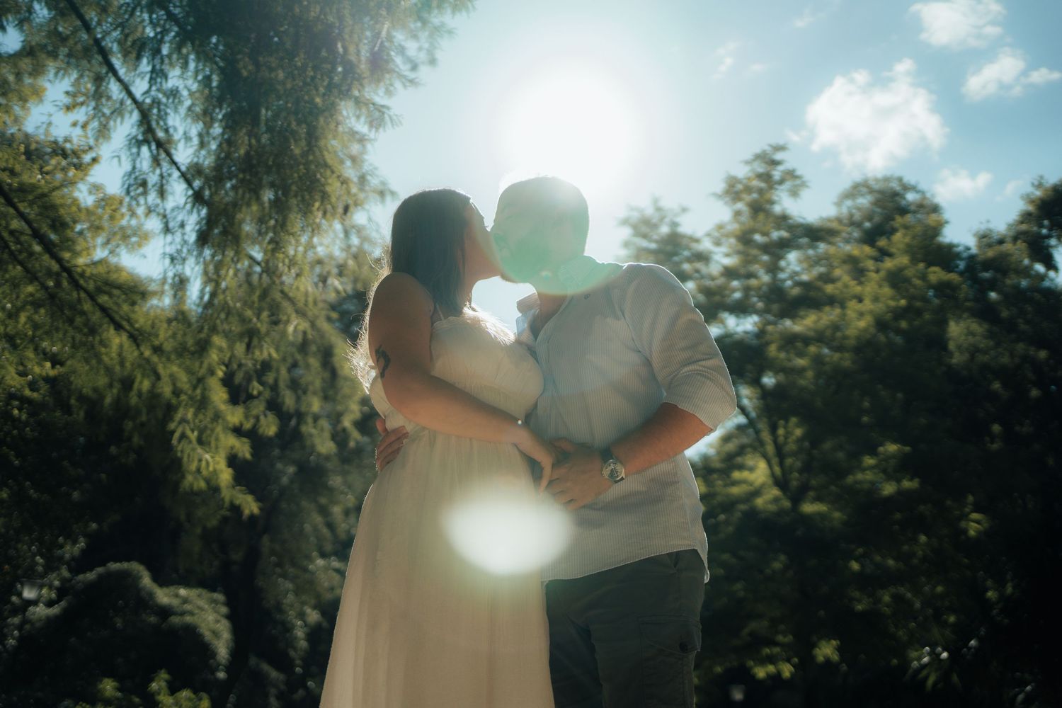 Couple kissing backlit by bright sun flare amid green trees, bride in flowing white dress.