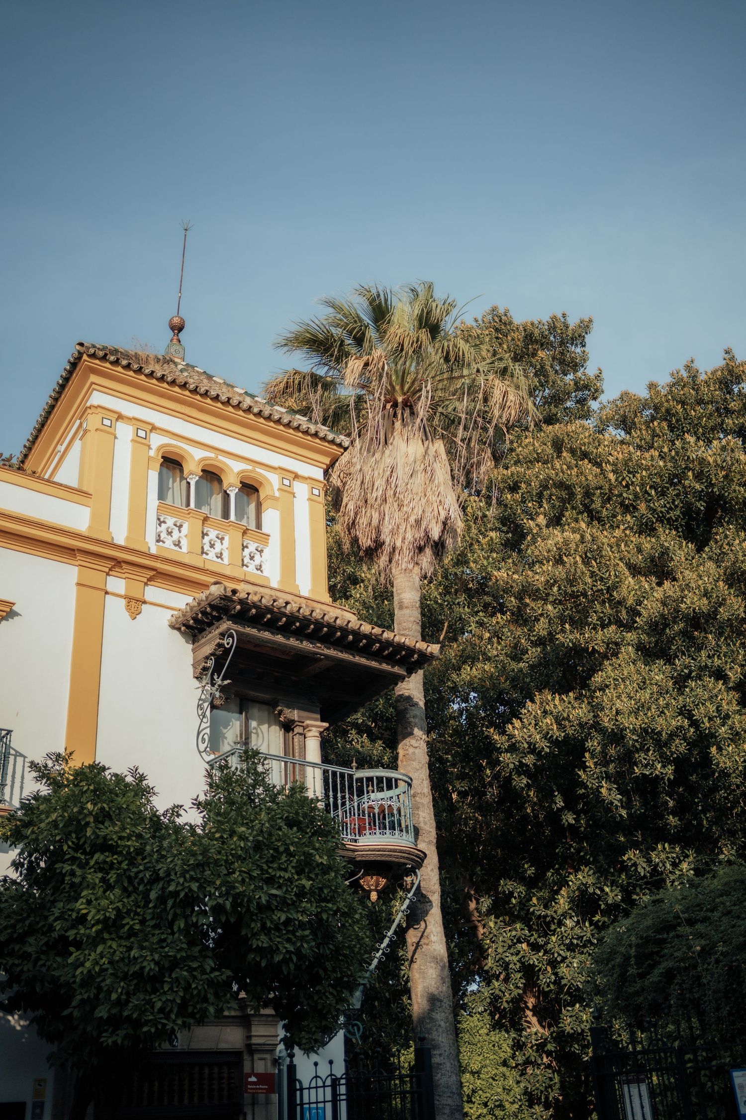 Andalusian style yellow and white building with palm trees and lush greenery under clear blue sky.