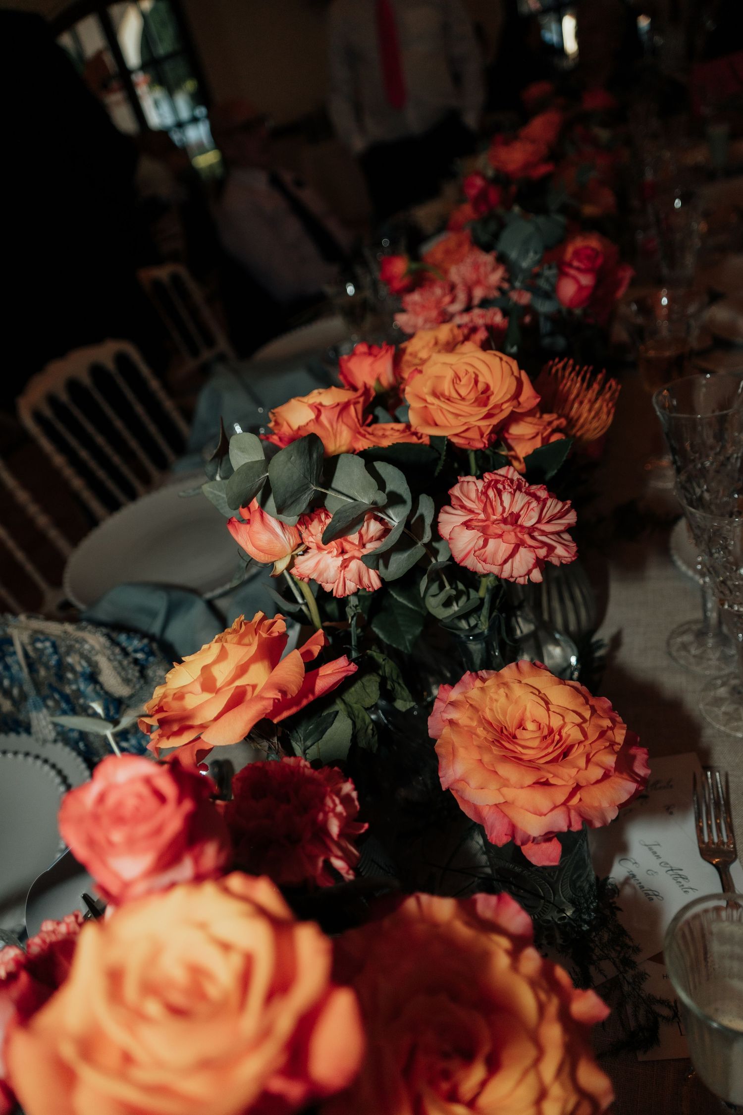 Elegant wedding table centerpiece featuring vibrant orange and pink roses with lush greenery on decorated table.