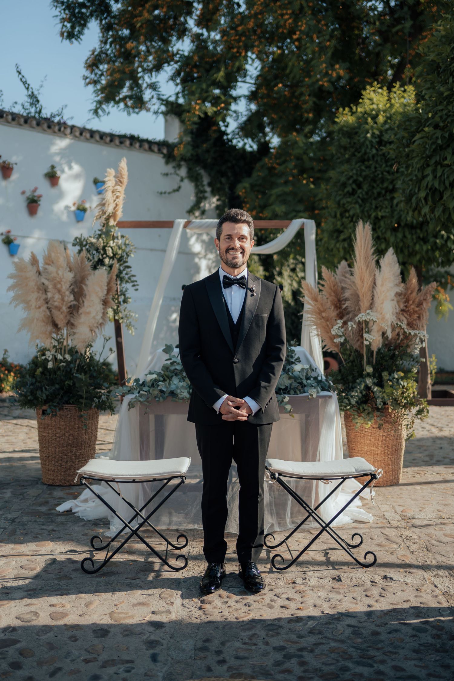 Smiling groom in black tuxedo stands before a floral arch with pampas grass at outdoor ceremony.