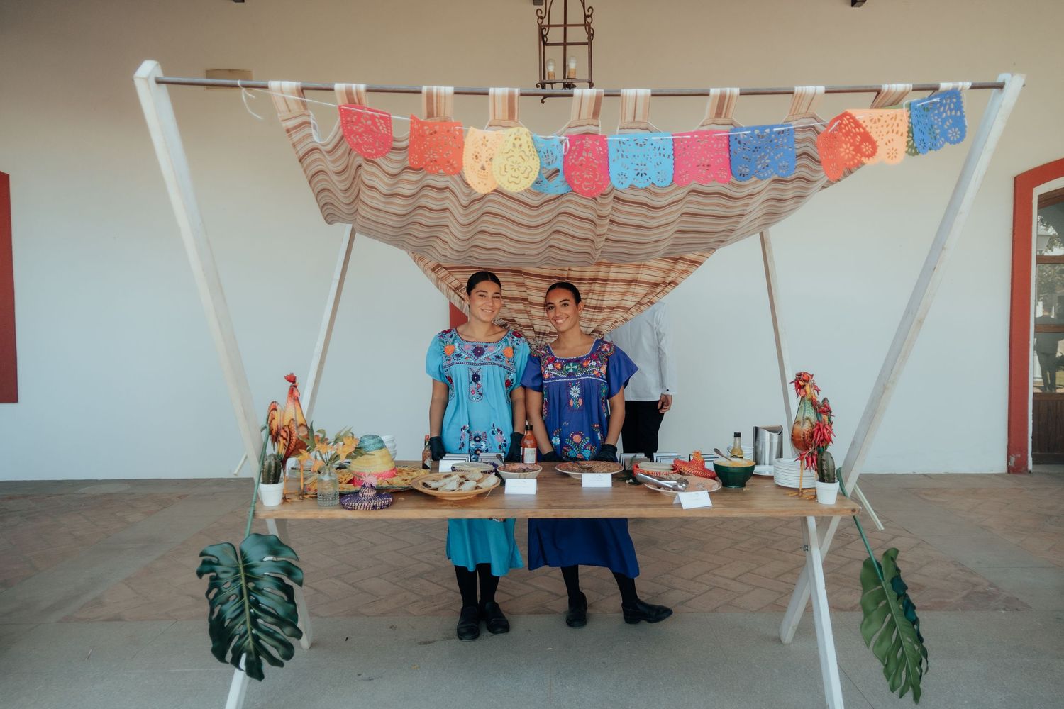 Two women in traditional Mexican attire stand behind a decorated market stall with papel picado banners.