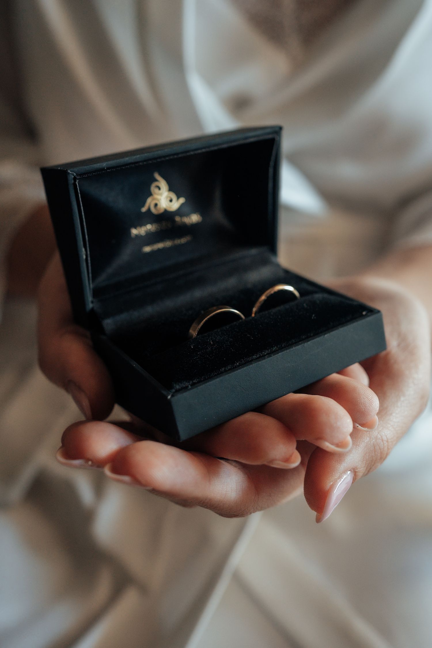 Close-up of hands holding an open black jewelry box containing two gold wedding bands.
