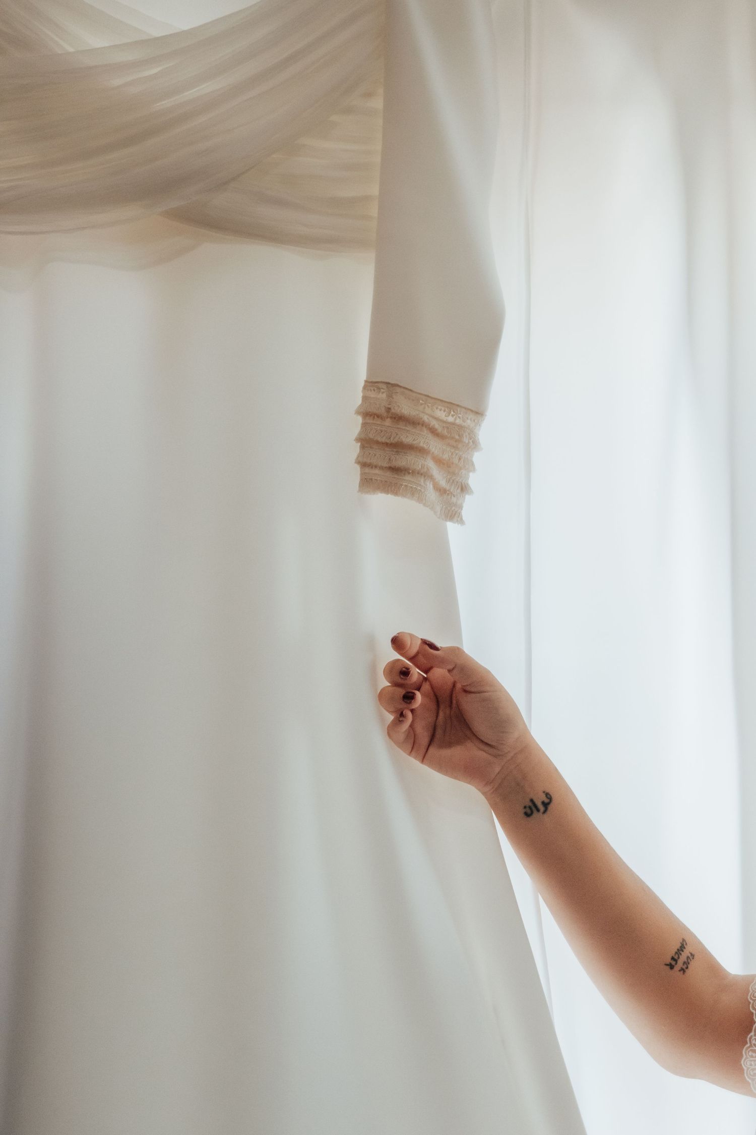 Bride's tattooed hand reaching up to touch her elegant white wedding dress hanging by a window.