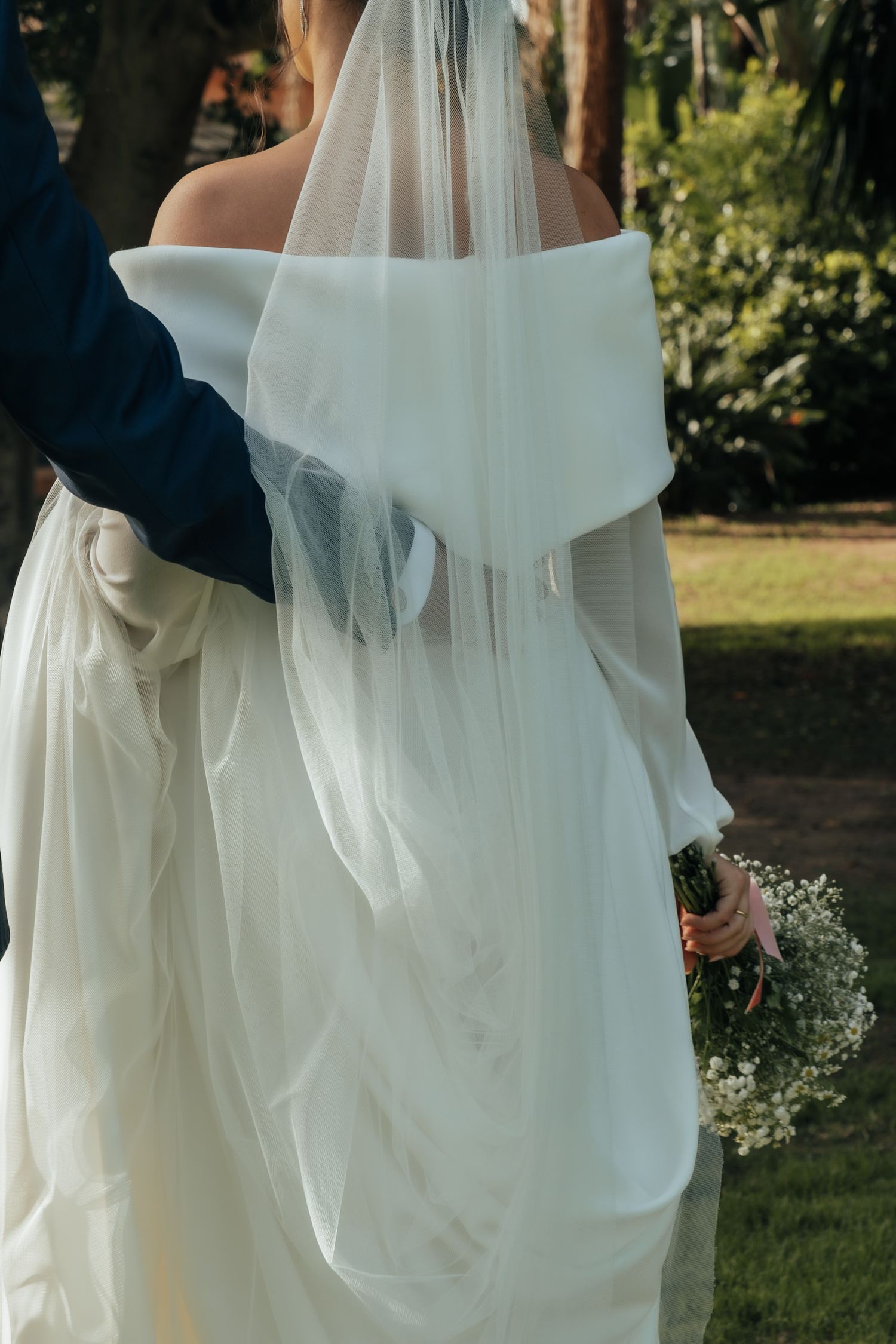 Back view of bride in off-shoulder white gown with veil, holding bouquet while groom embraces her outdoors.