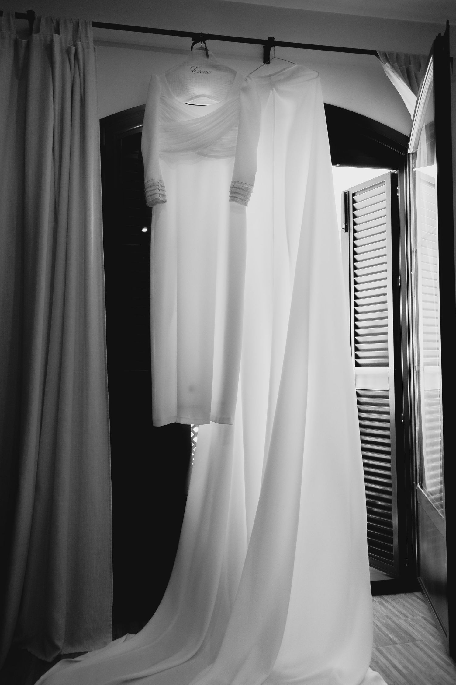 Black and white photo of wedding dress and veil hanging gracefully near arched window.