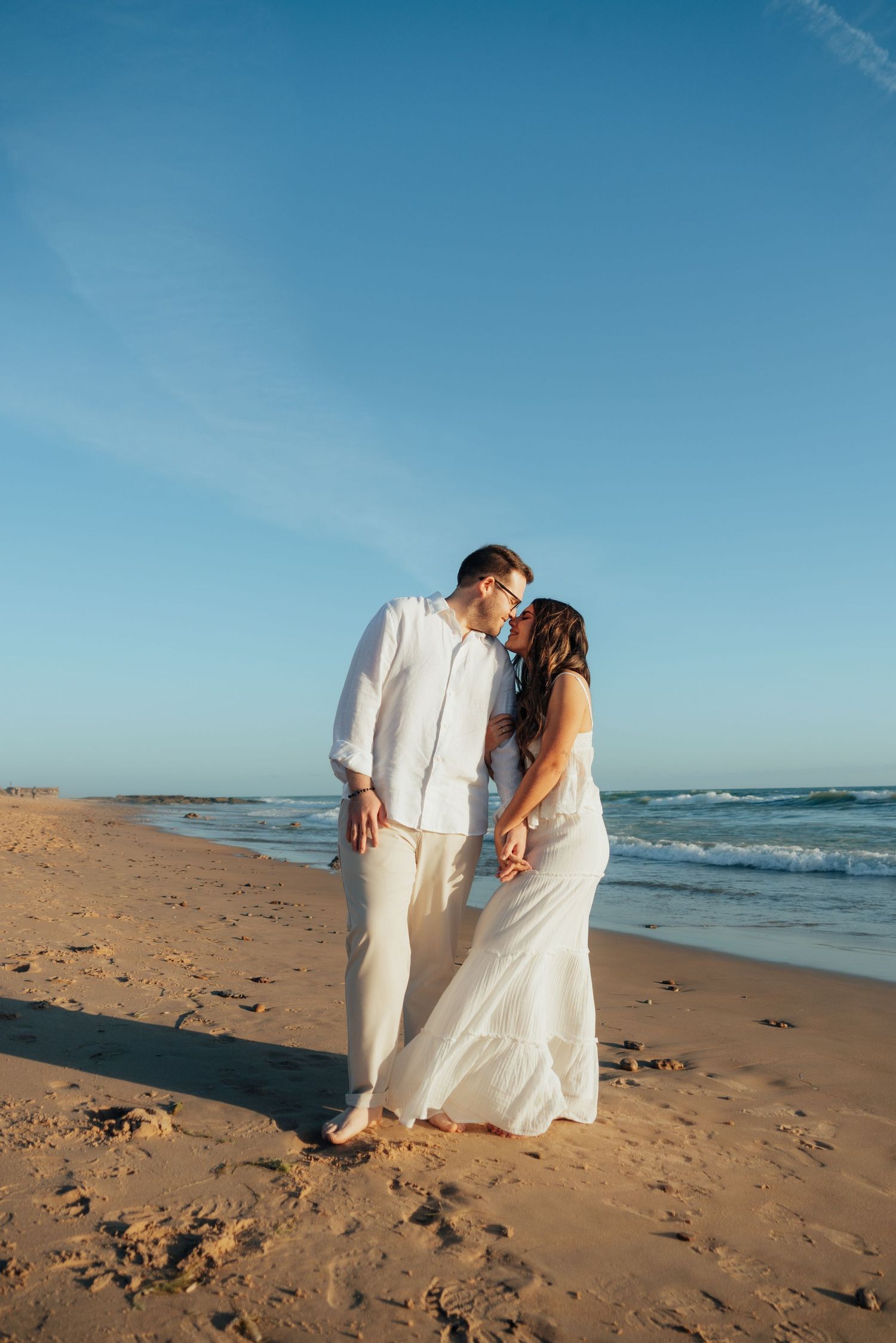 Couple in white outfits sharing a romantic kiss on a sandy beach at sunset with blue sky and ocean waves.