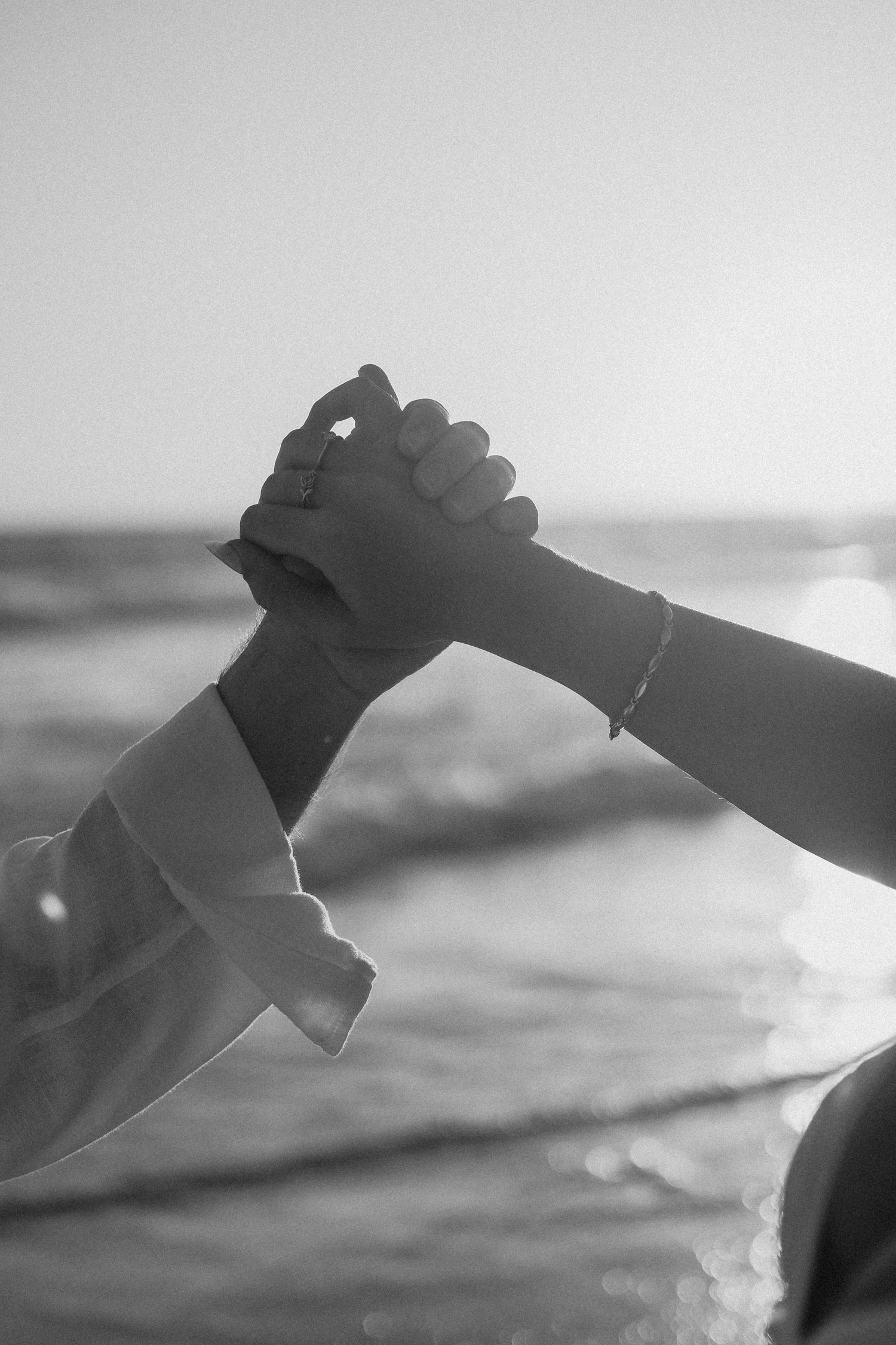 Black and white photo of two people holding hands at the beach, with ocean waves in the background.