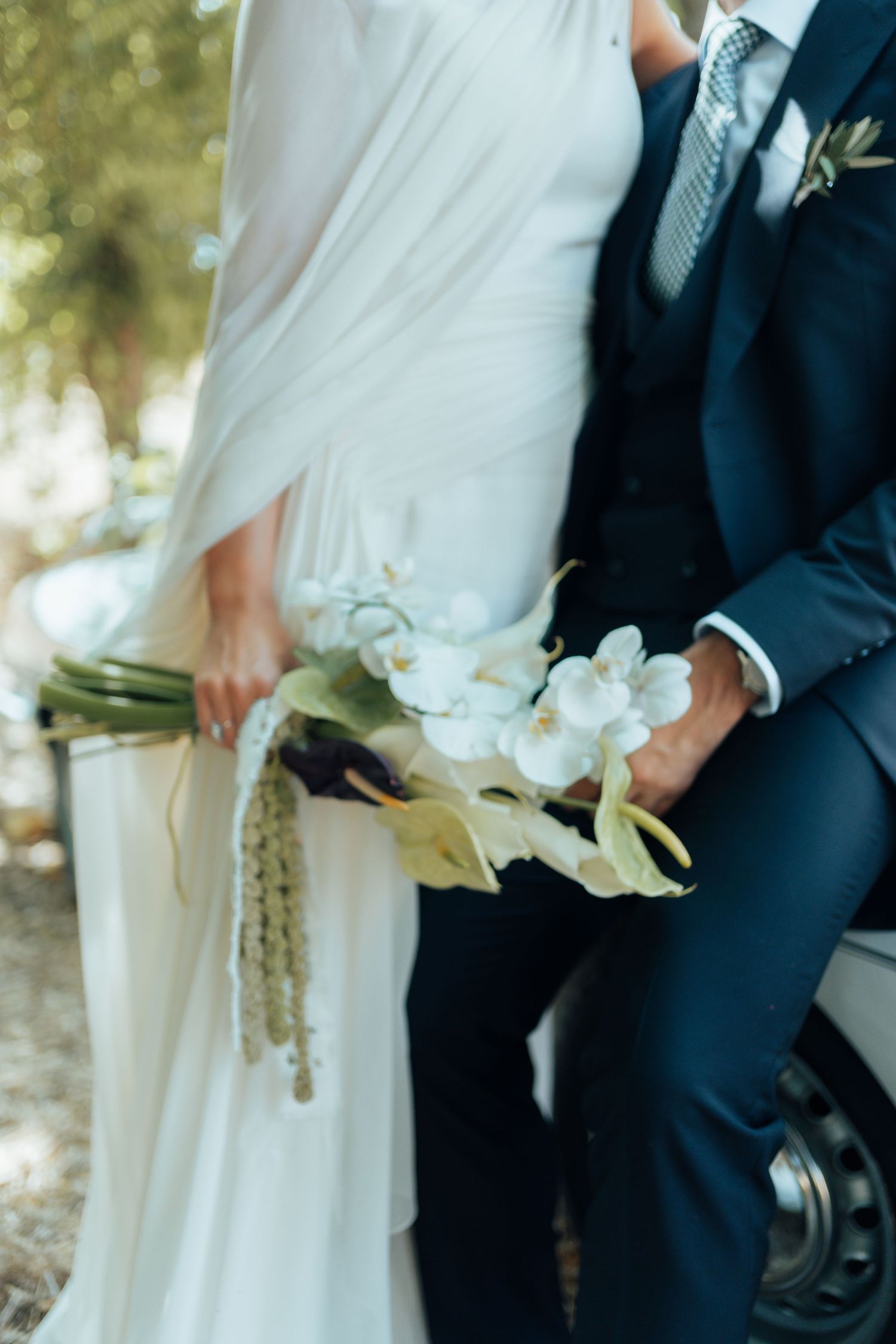 Bride in white gown and groom in navy suit holding elegant white orchid wedding bouquet outdoors.
