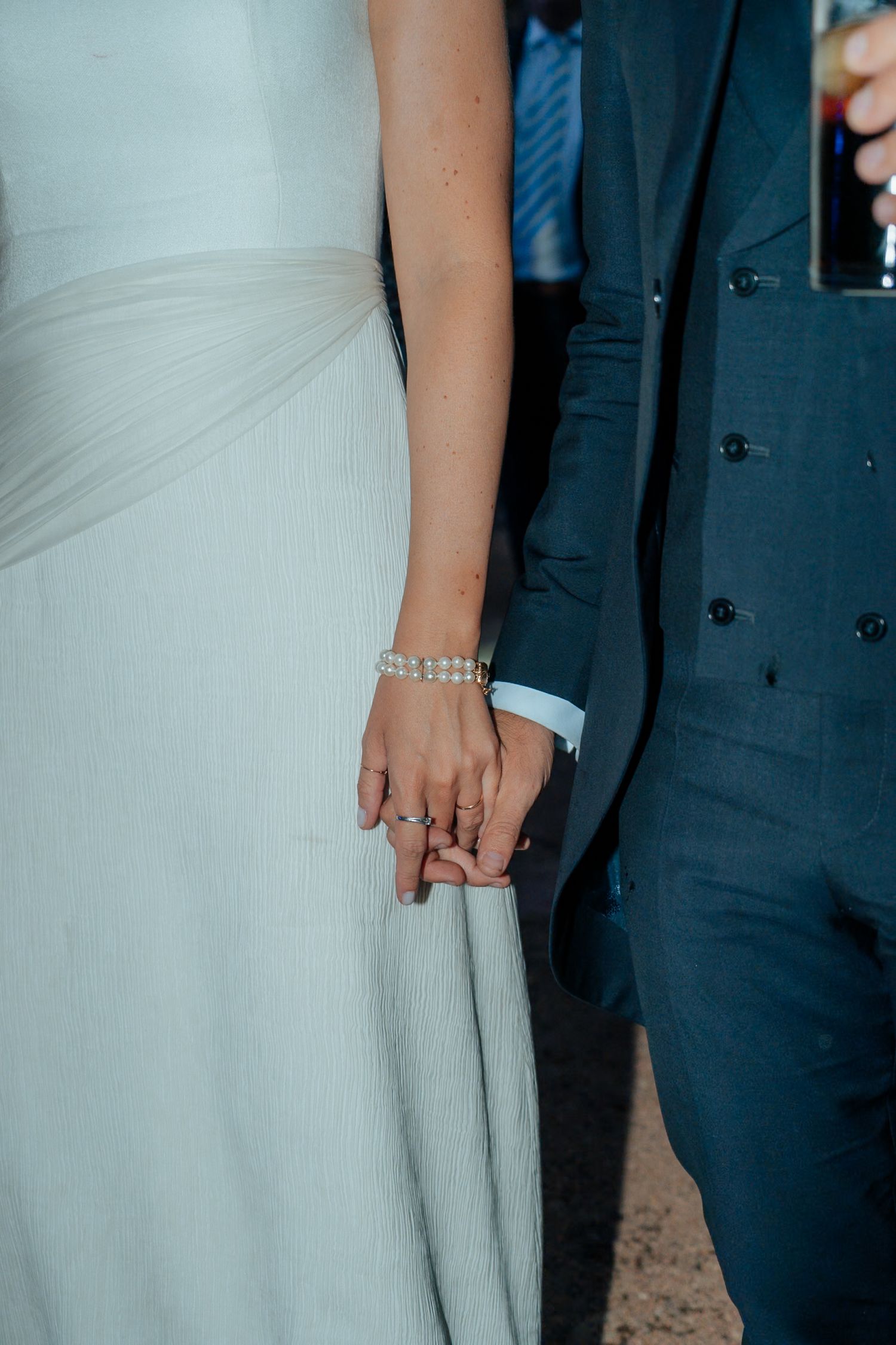 Close-up of bride and groom holding hands, bride wearing pearl bracelet and wedding ring.