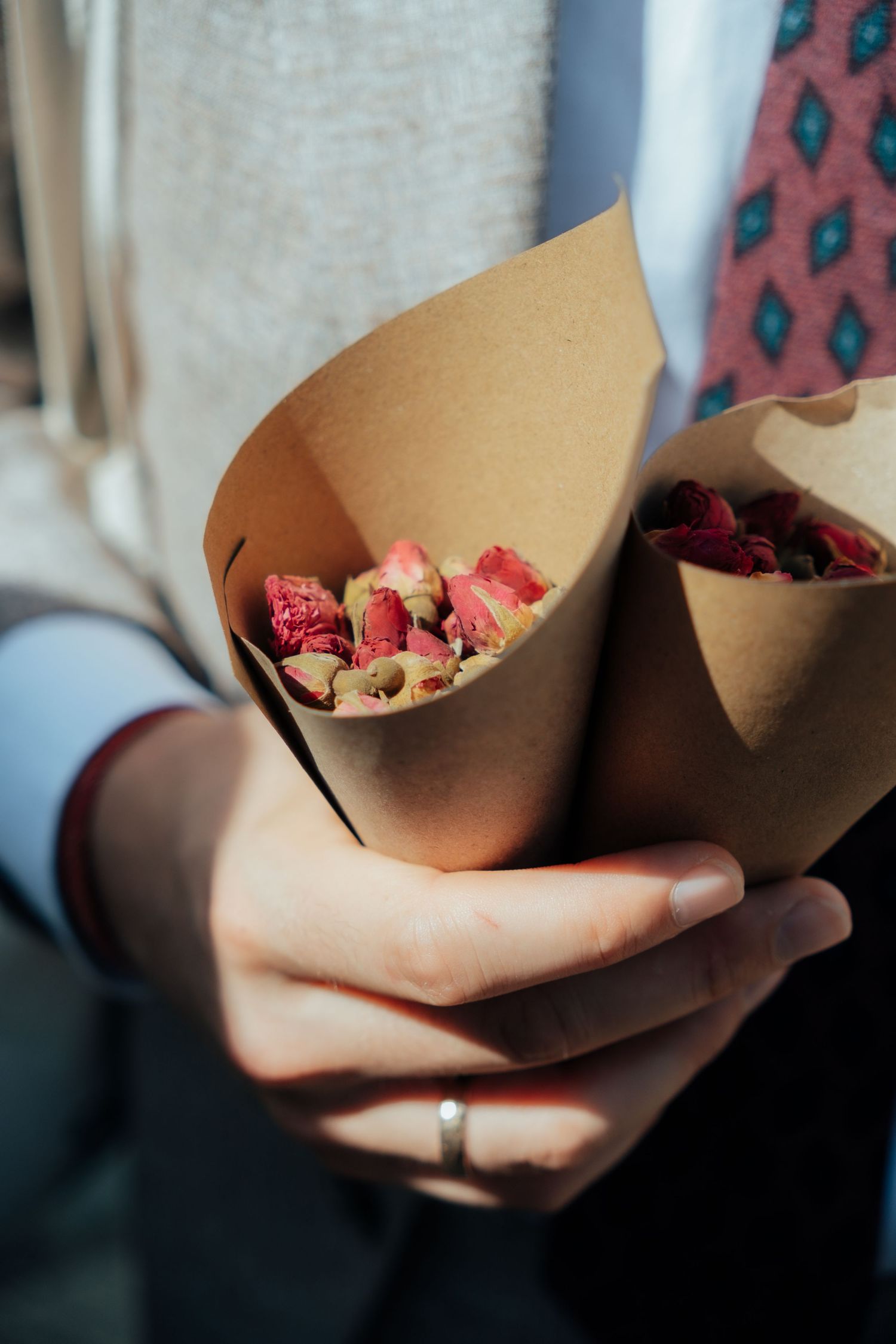 Groom holding two kraft paper cones filled with dried rose petals as wedding confetti outdoors.