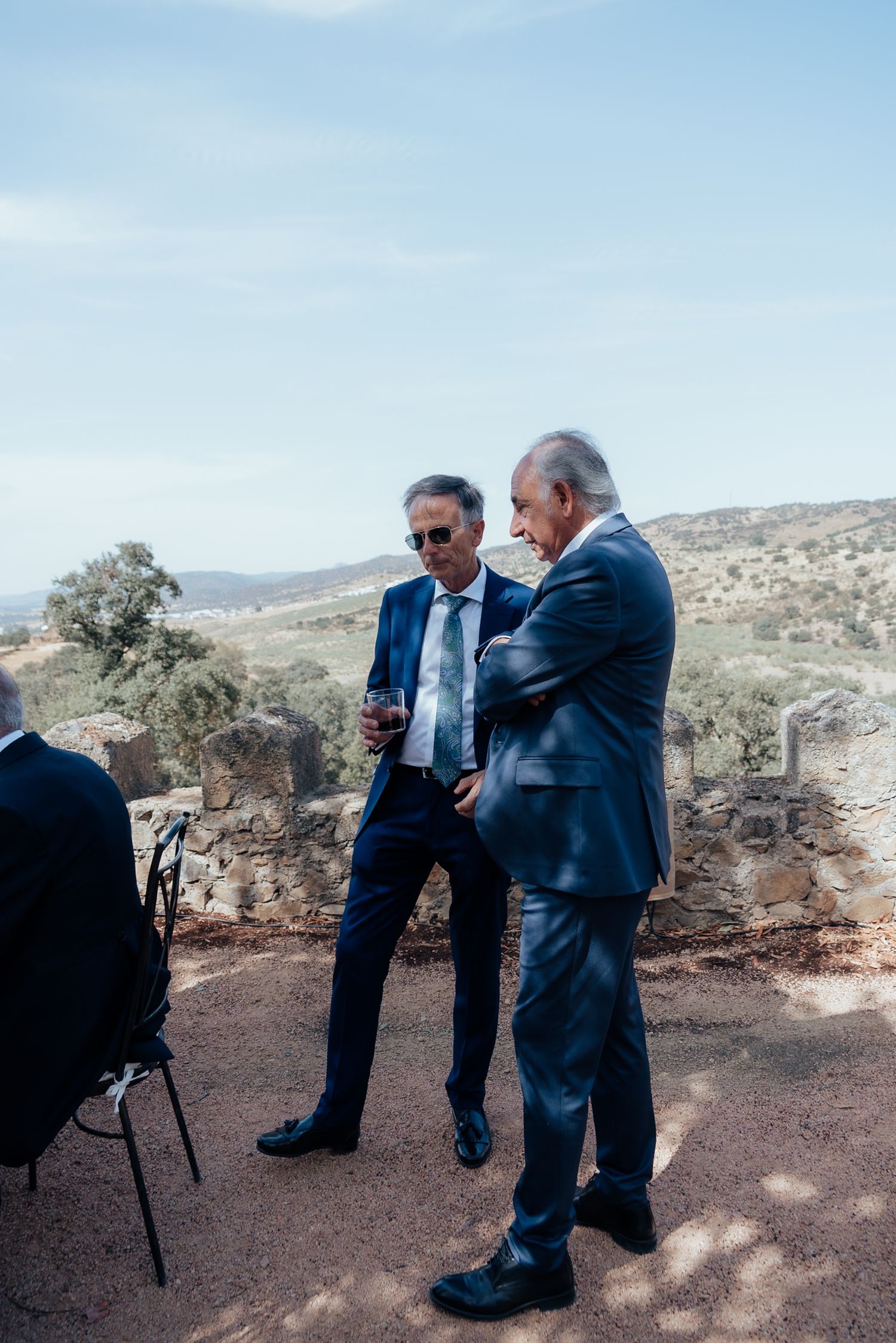 Two elegant older men in navy suits conversing at outdoor wedding with scenic landscape view.