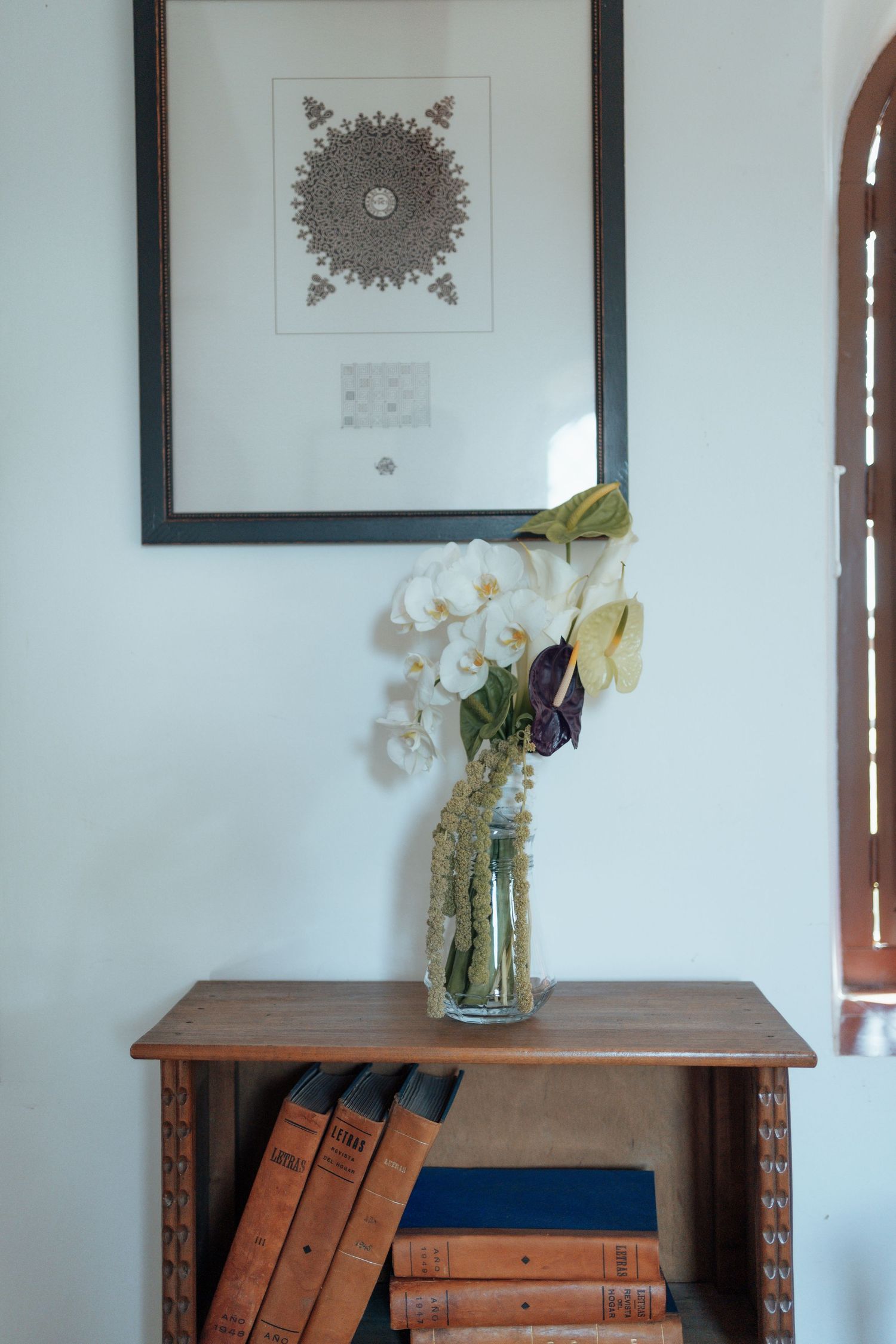White orchid bouquet in glass vase on wooden bookshelf beneath framed mandala artwork.