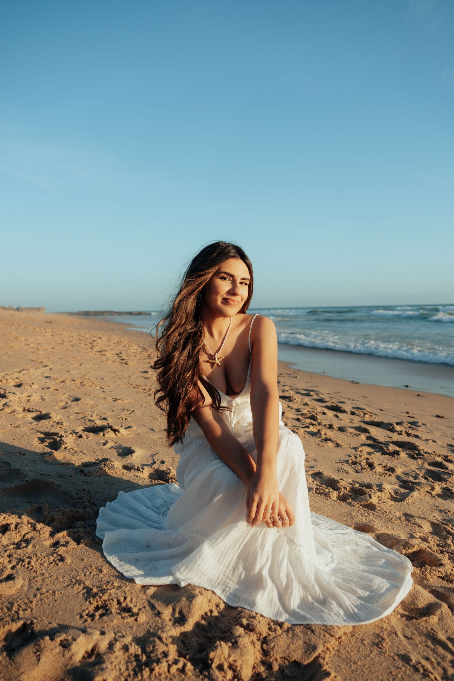 Young woman in white dress sitting on sandy beach with ocean waves and blue sky backdrop.