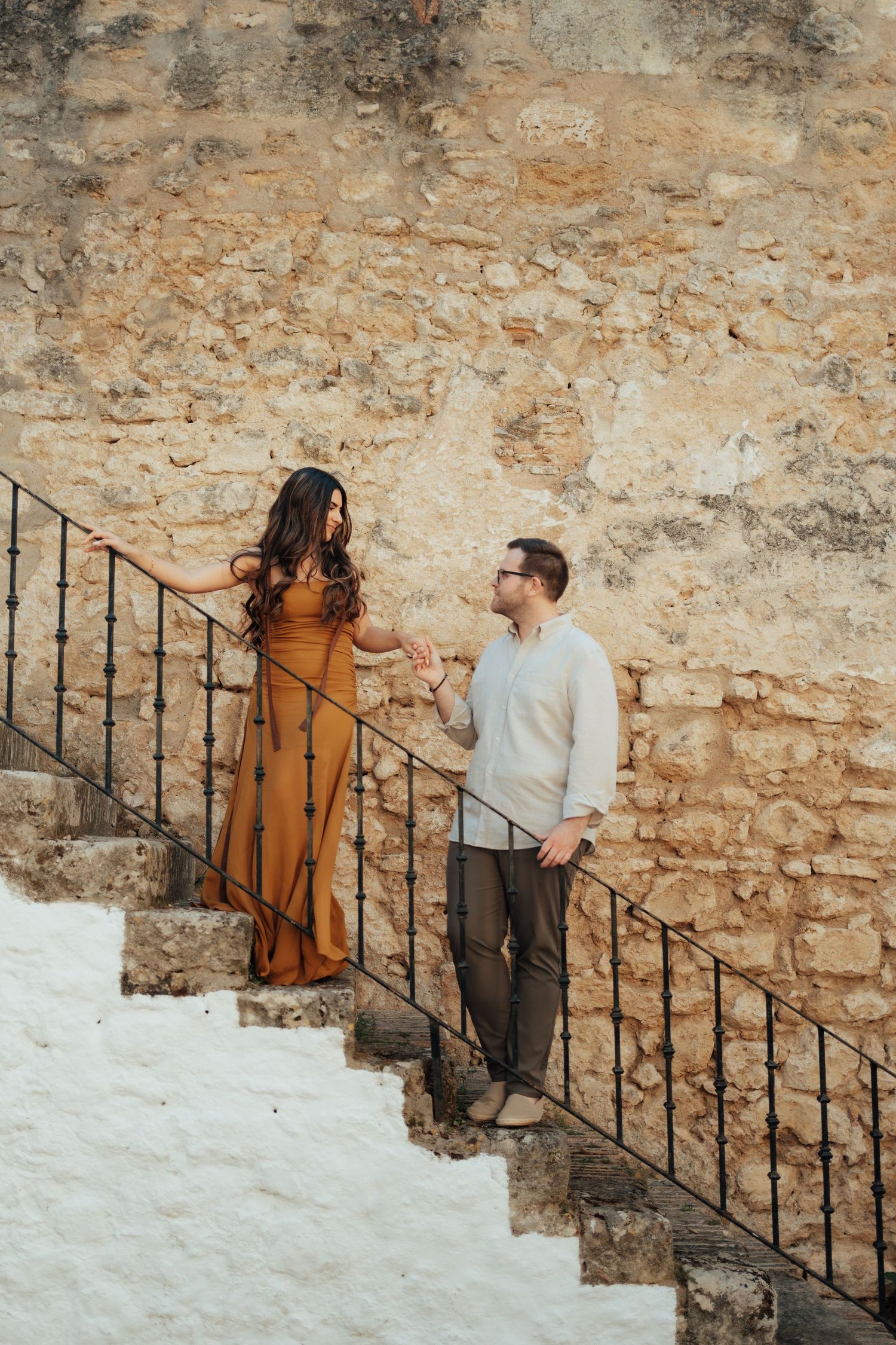 Couple holding hands on stone steps by rustic wall, woman in orange dress, man in white sweater.