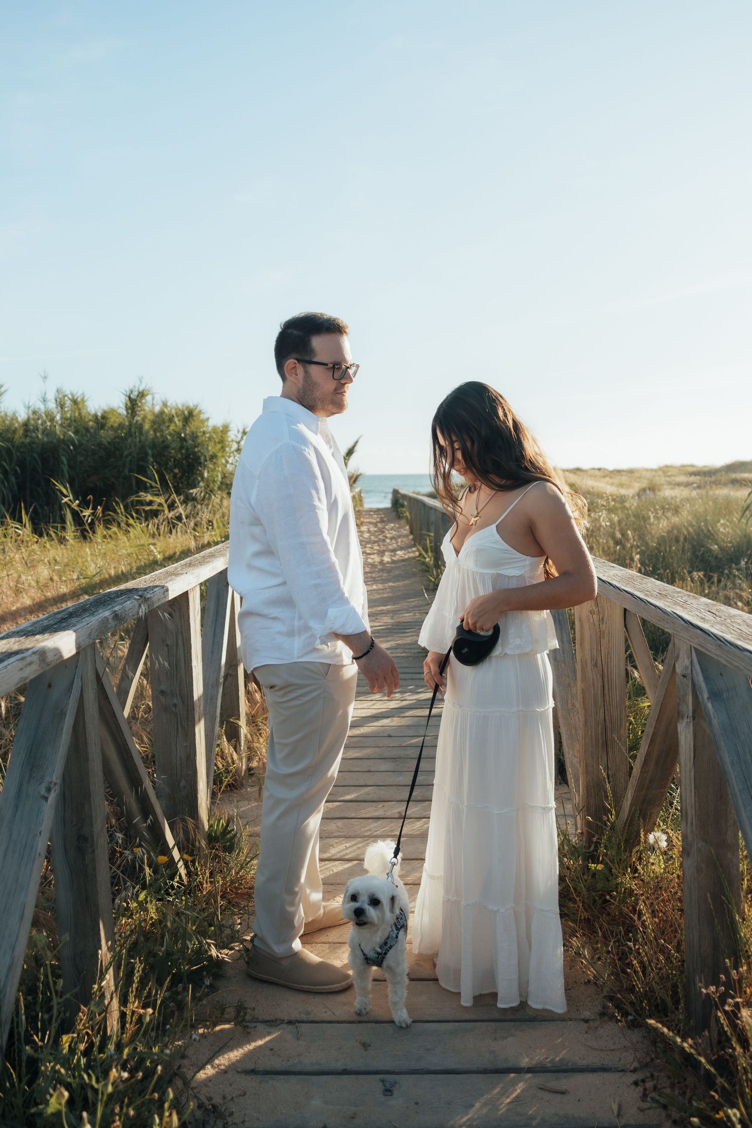 Couple in white outfits holding hands with small dog on wooden beach boardwalk path surrounded by dunes.