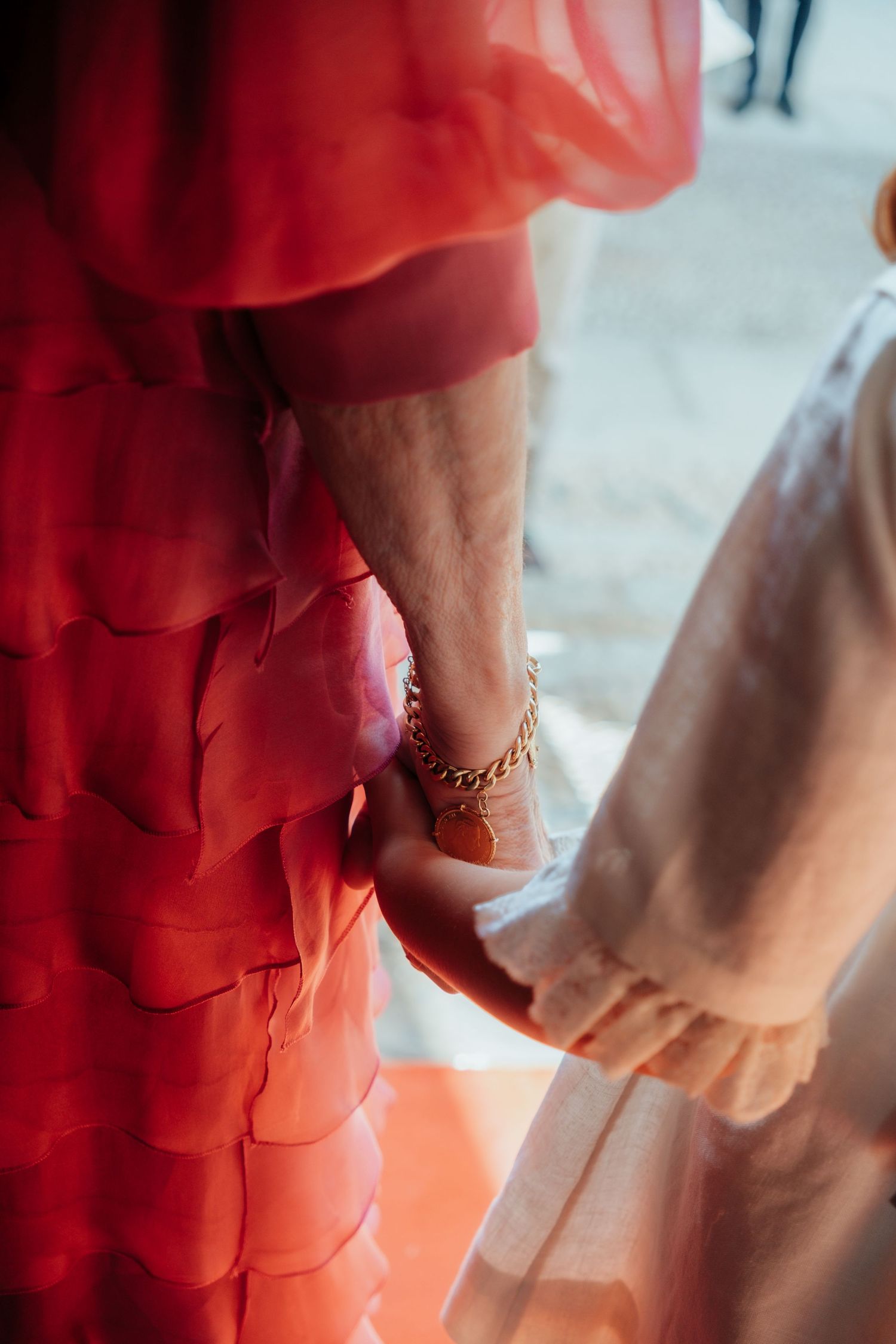 Close-up of hands holding with gold chain bracelet, red ruffled dress and white outfit at wedding.