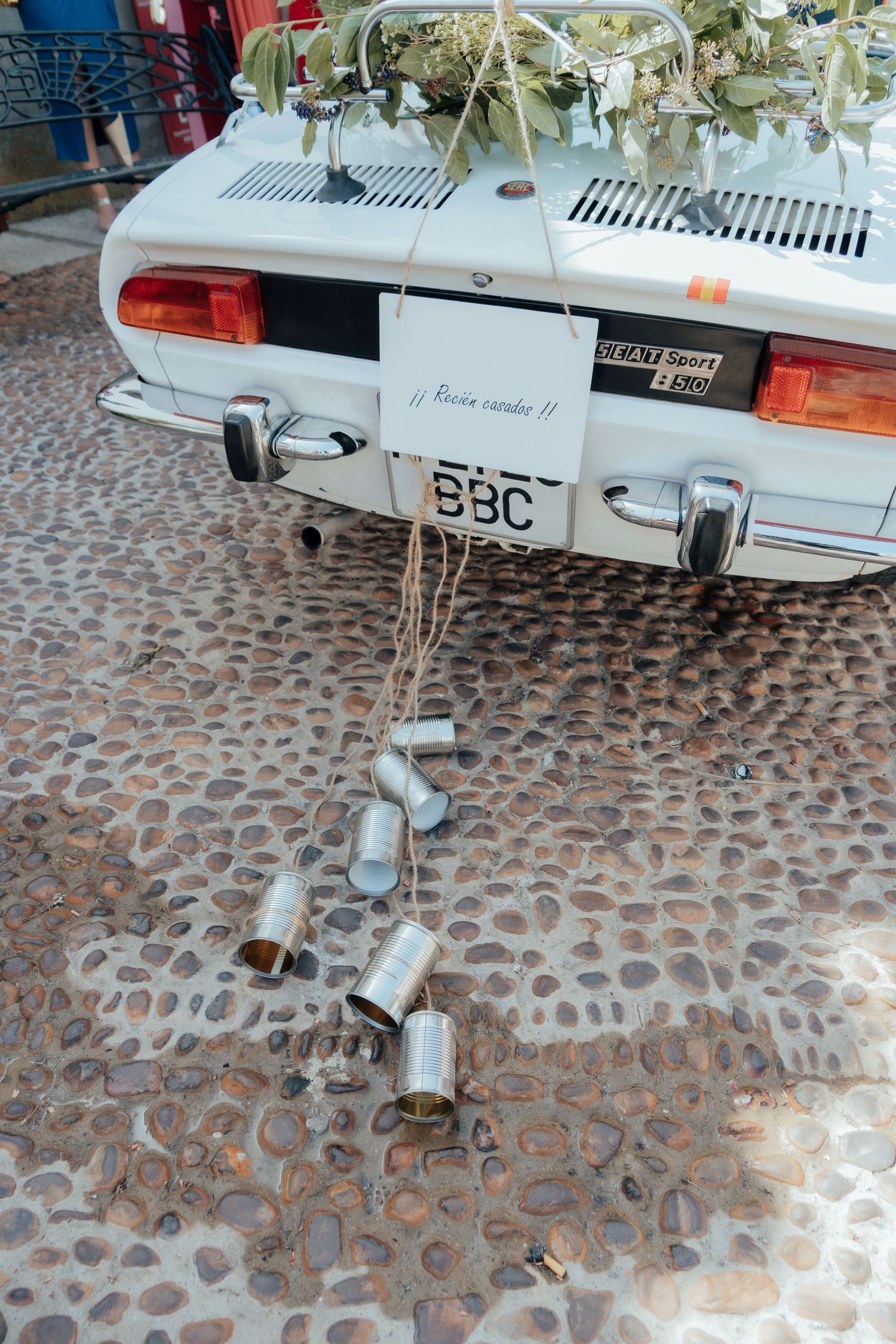 Tin cans tied to the back of a white vintage wedding car with BBC license plate on cobblestone ground.