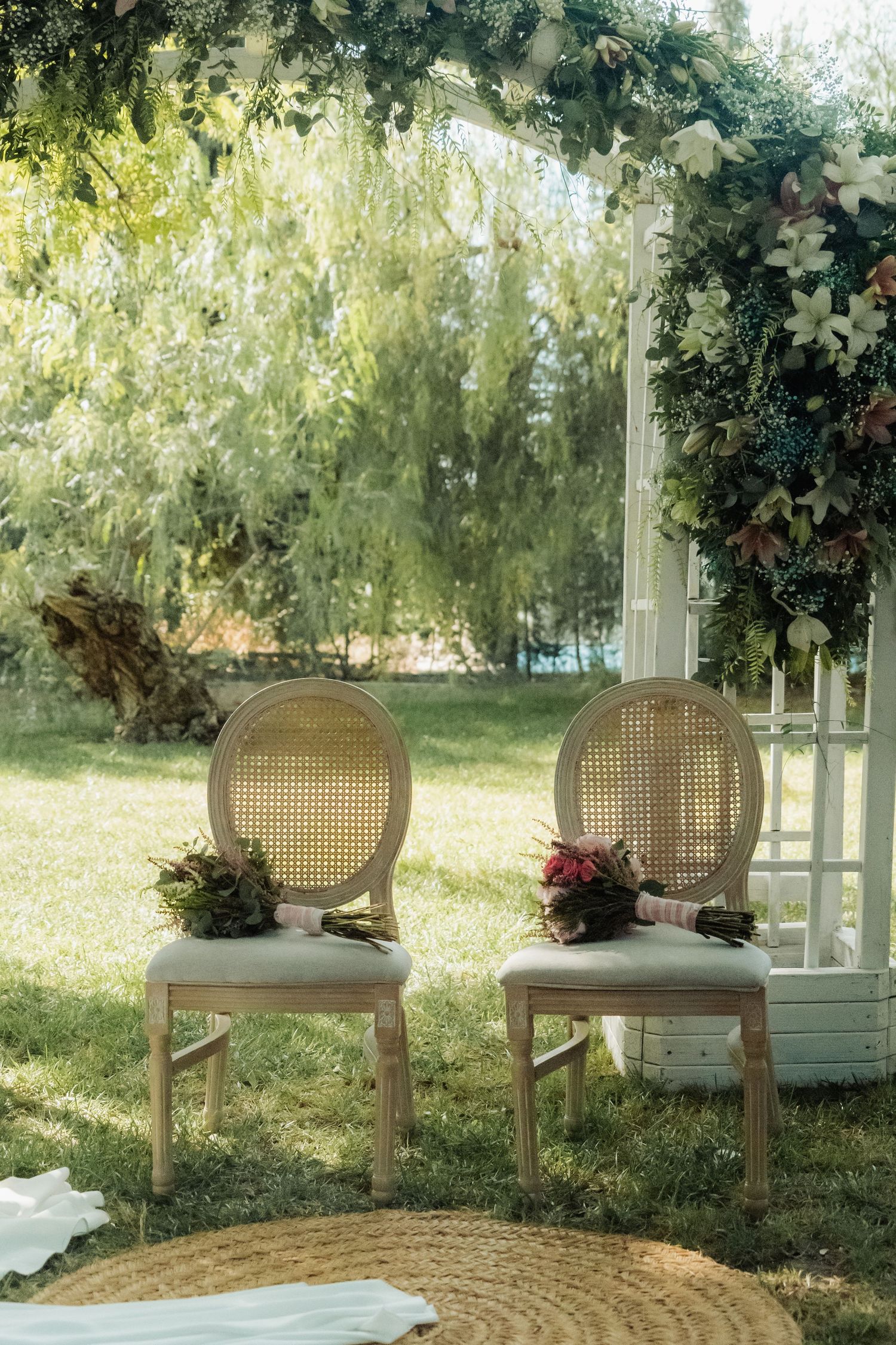 Two rattan chairs with bridal bouquets beneath floral arch at outdoor wedding altar.