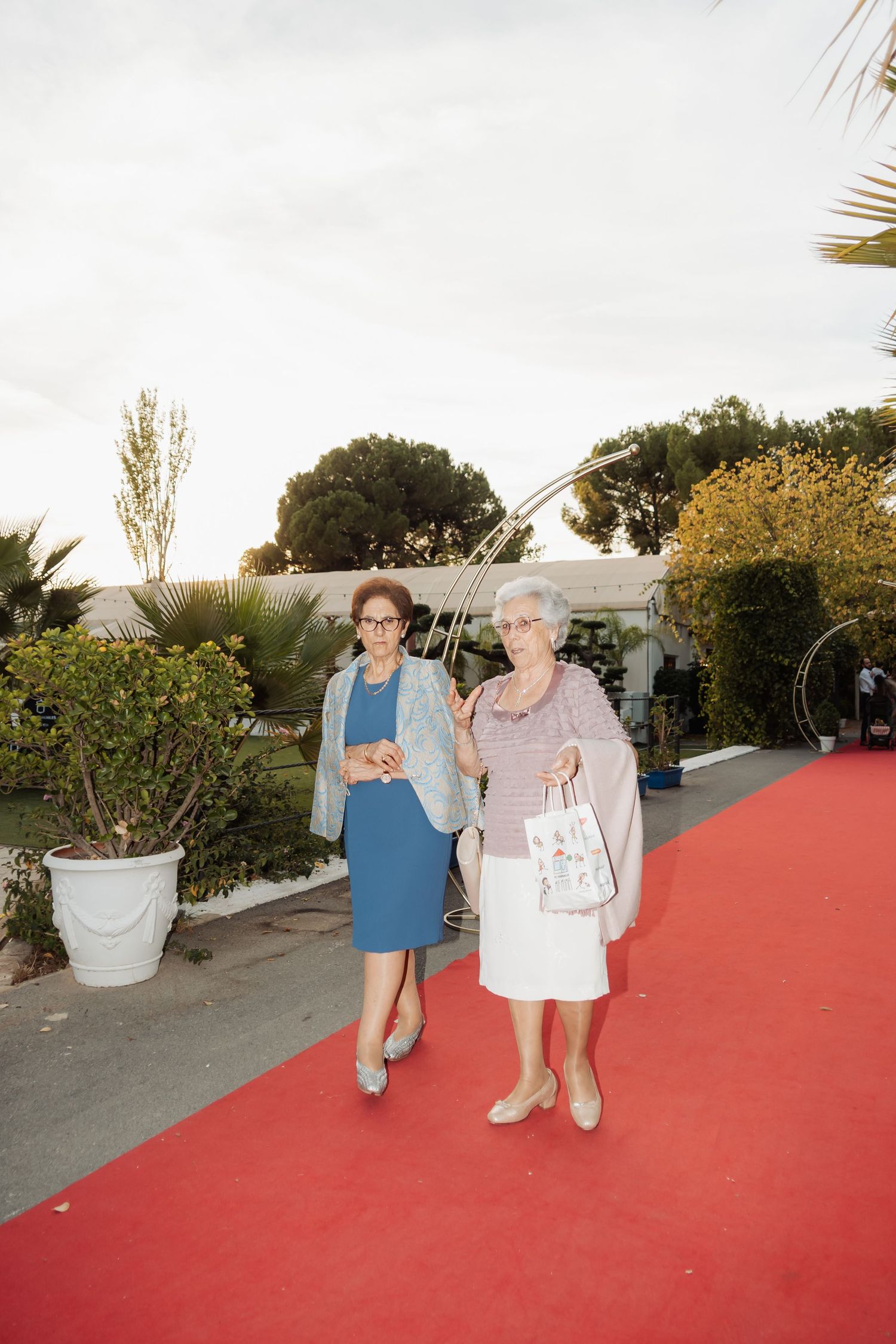 Two elderly women in formal attire walking together on a red carpet at an outdoor event.