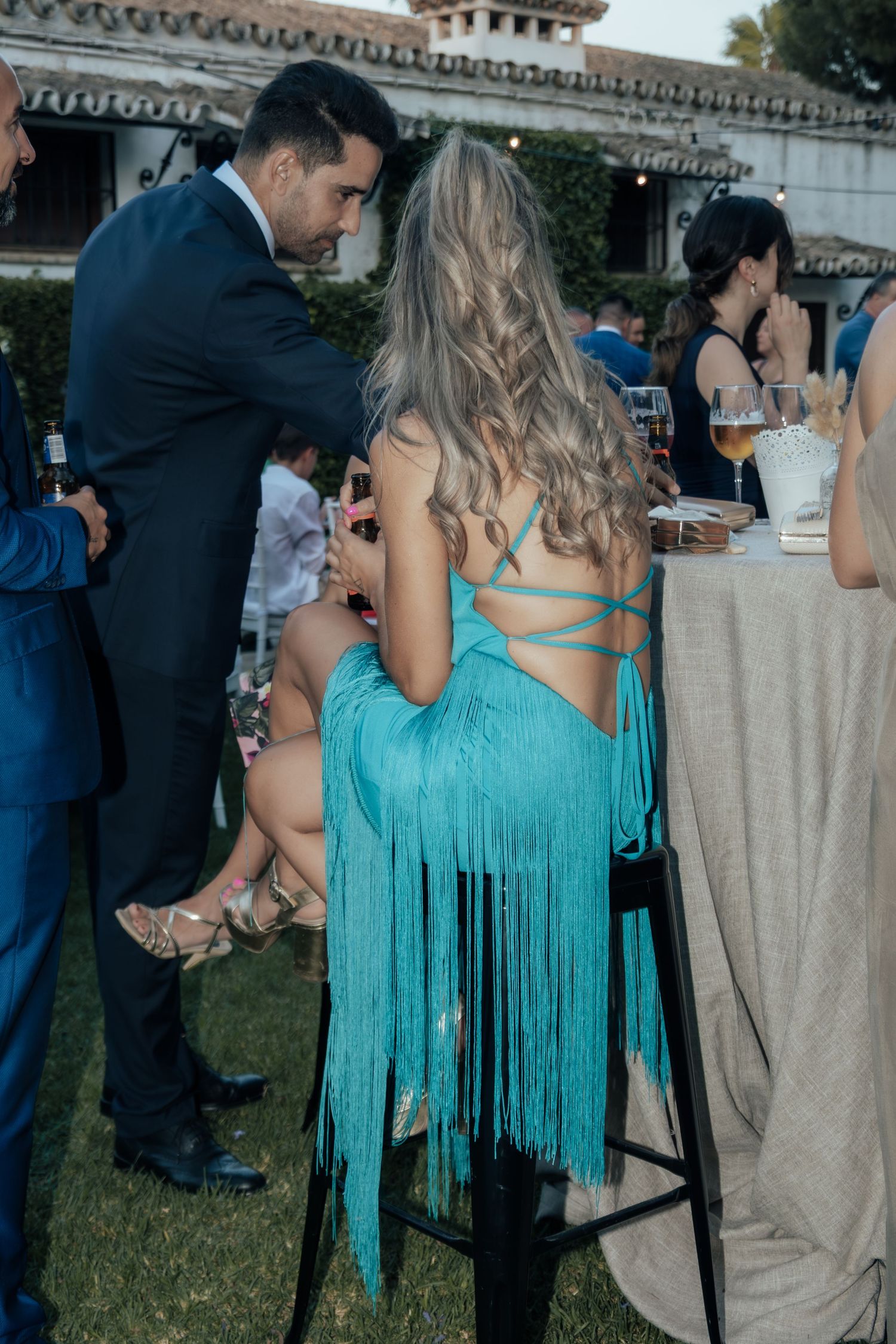 Wedding guest in turquoise fringed backless dress seated on bar stool at outdoor evening reception.