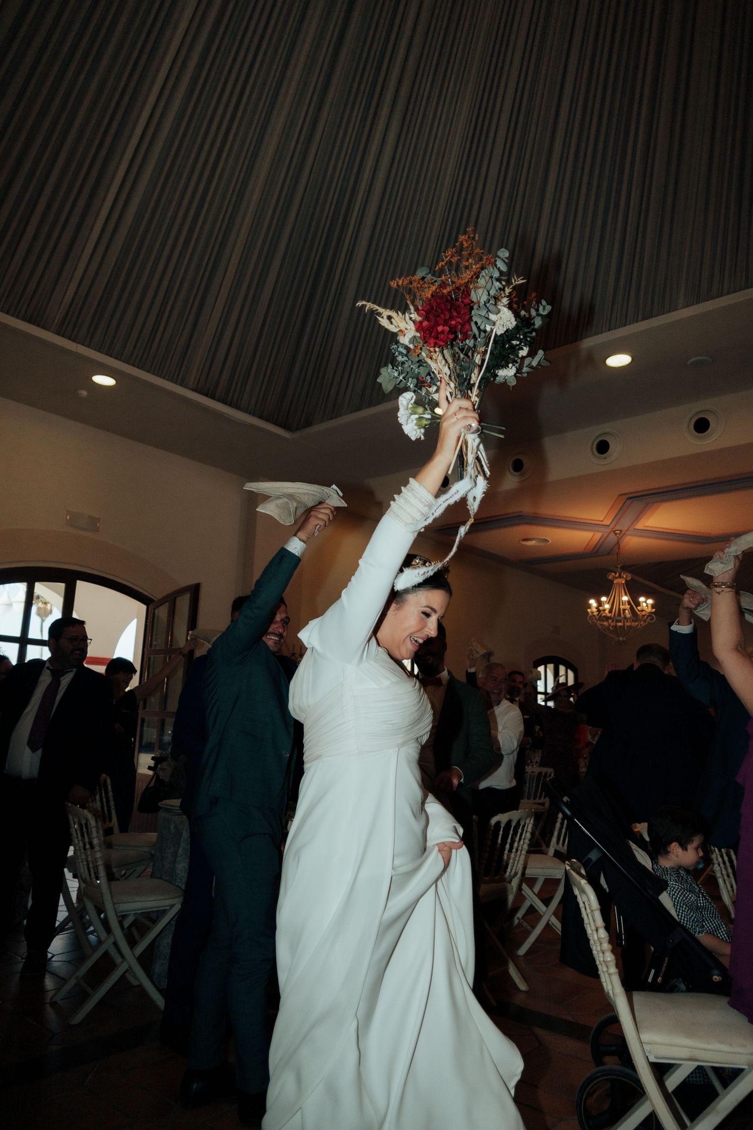 Joyful bride in white long-sleeve gown raises colorful bouquet while celebrating at indoor wedding reception.
