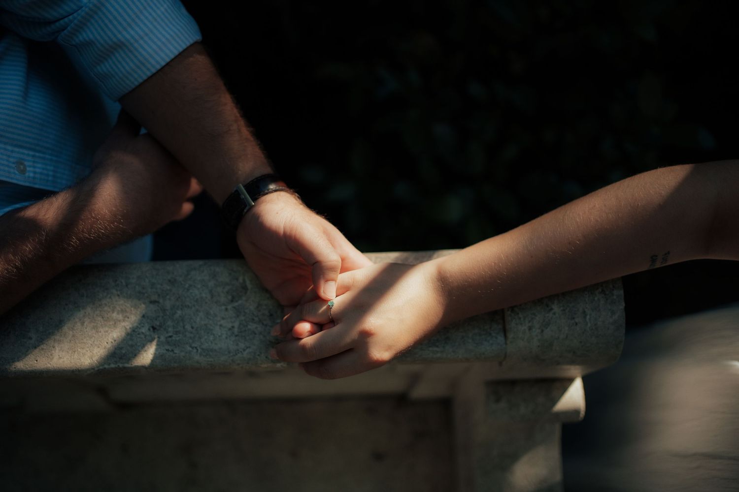 A close-up of a couple holding hands on a stone railing, with soft sunlight illuminating their intertwined fingers.