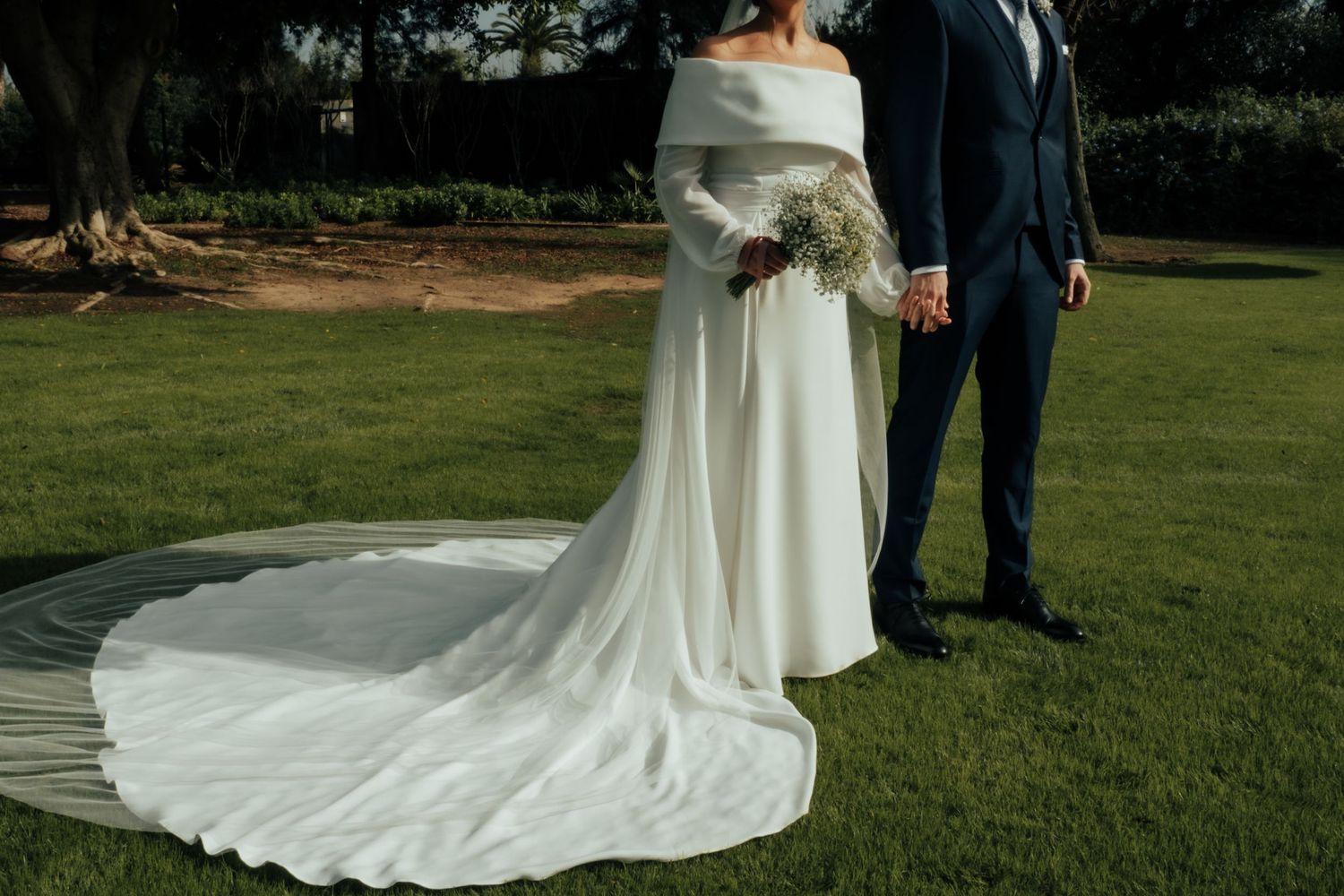 Bride in elegant off-shoulder white gown with cathedral train holds bouquet while groom stands beside her.