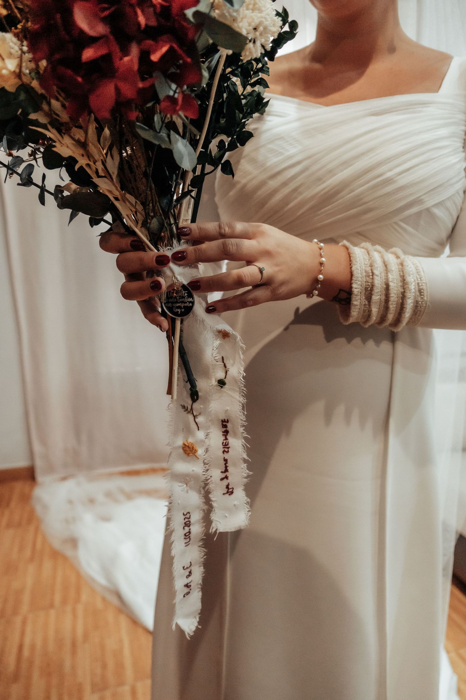 Bride in white long-sleeve gown holding bouquet with decorative ribbon and floral arrangement.