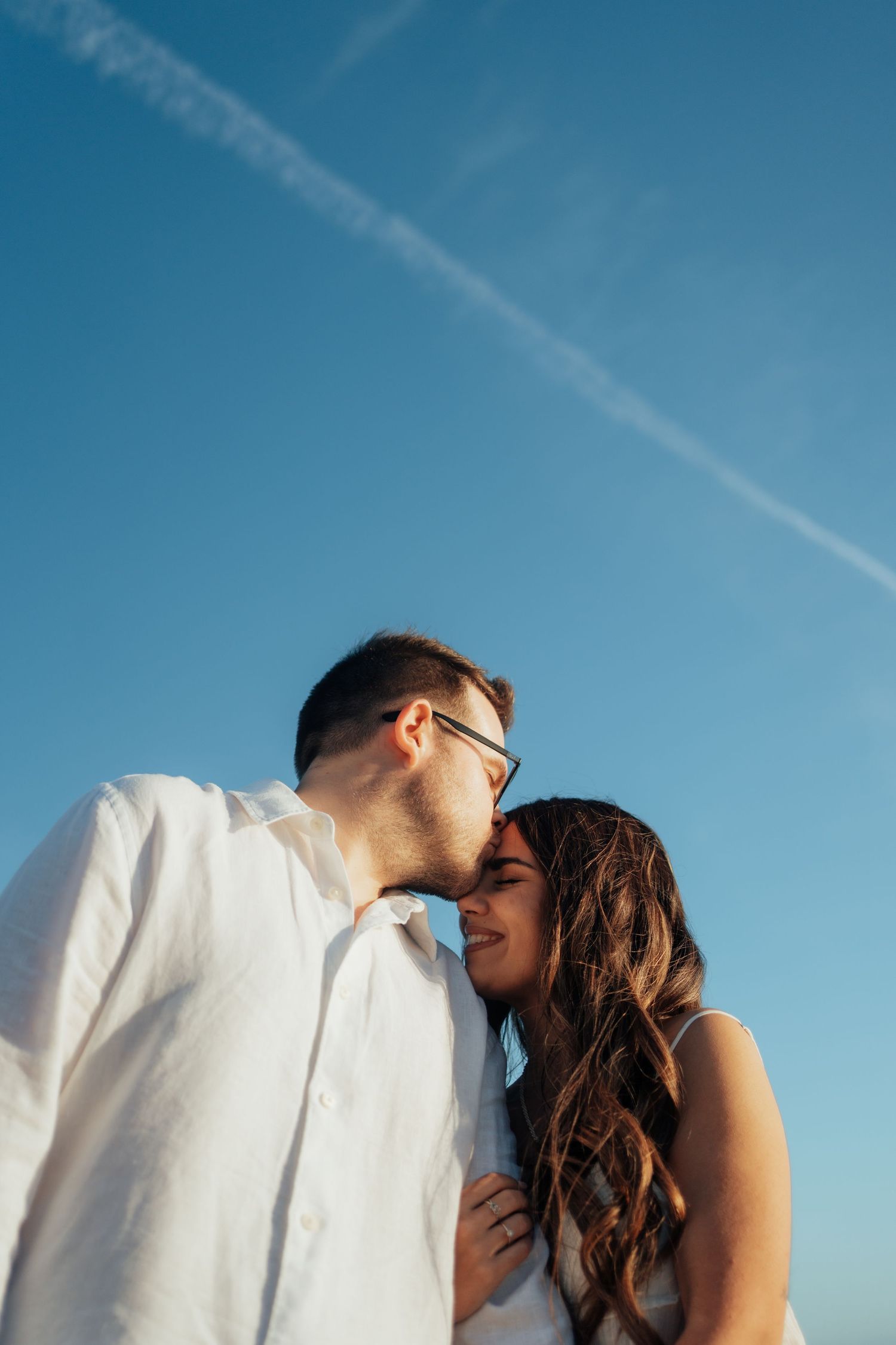 Romantic couple sharing a tender moment under a clear blue sky, man in white shirt kissing woman's head.