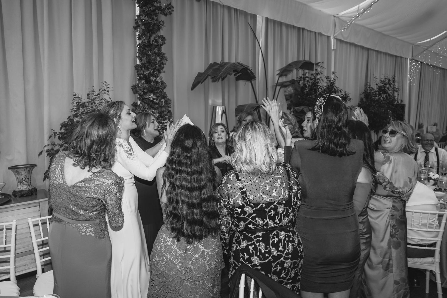 Black and white photo of wedding guests dancing together under floral arch at reception.