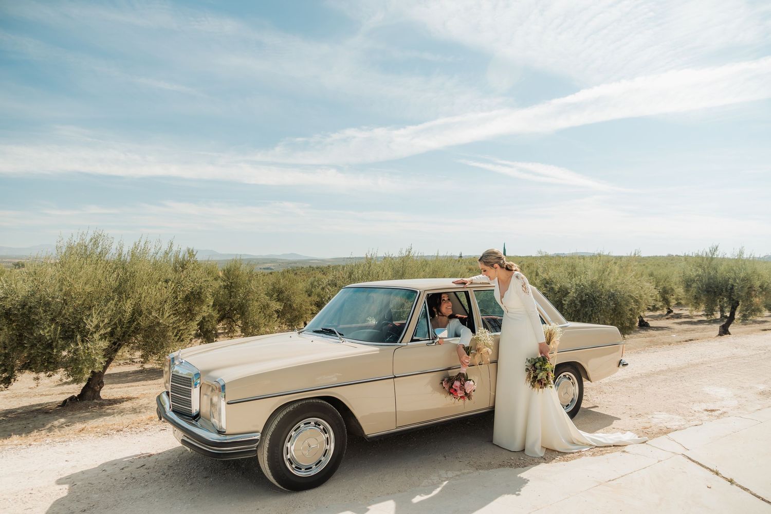 Bride in white gown poses beside vintage beige Mercedes in scenic olive grove on wedding day.