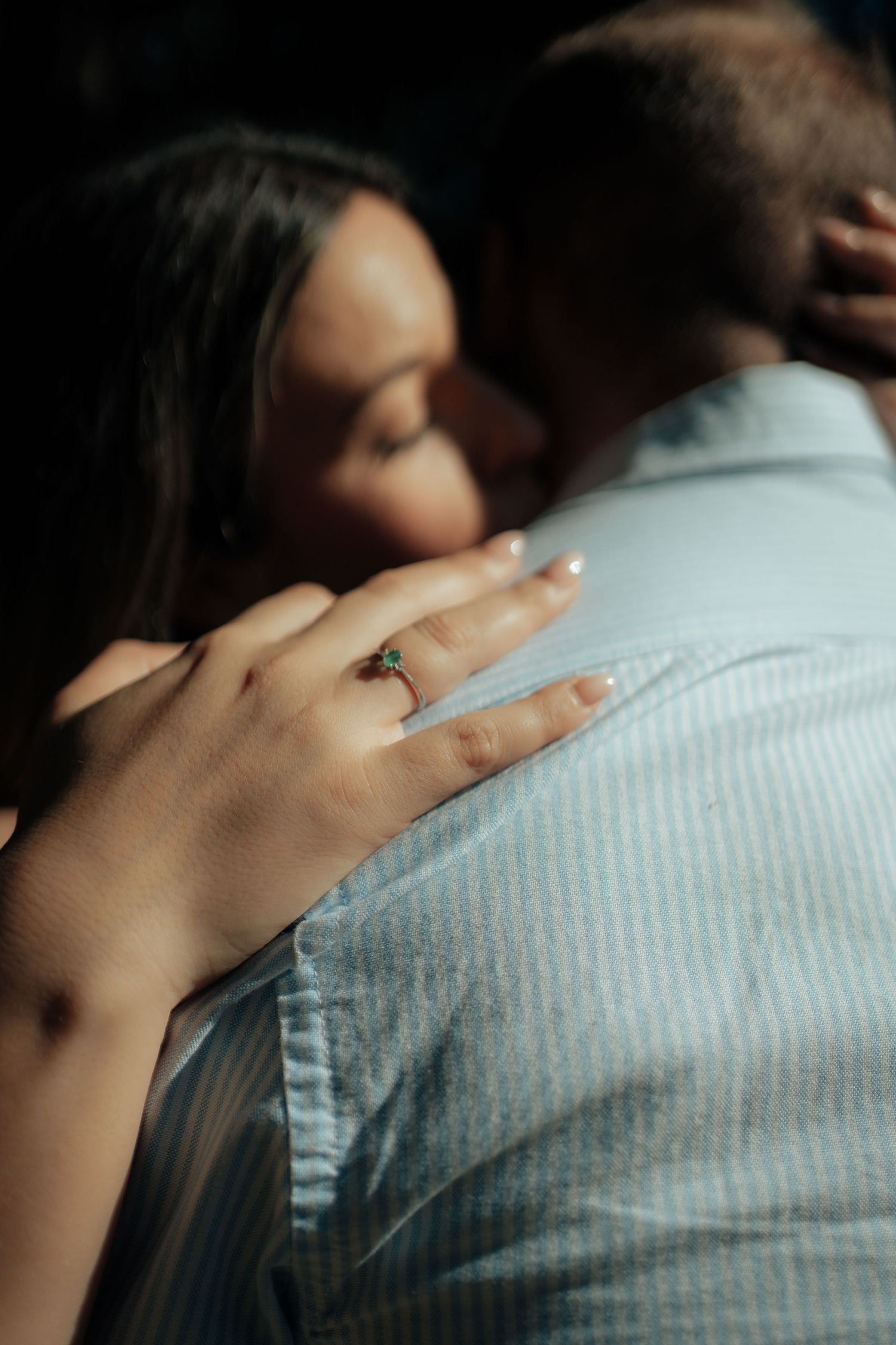Close-up of womans hand with green gemstone engagement ring resting on mans light blue shirt.