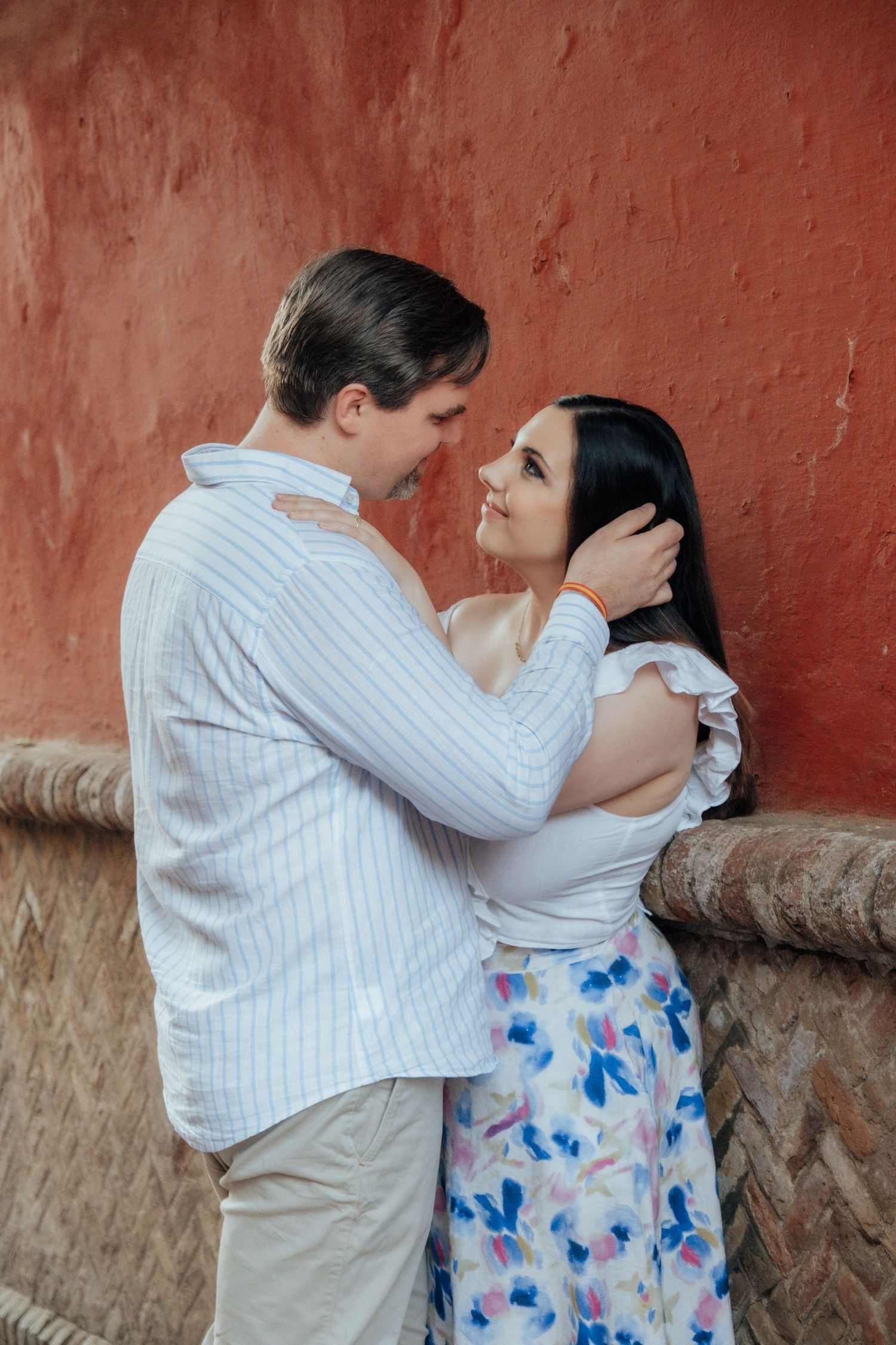 Romantic couple embracing and smiling at each other against a terracotta red wall in Seville.