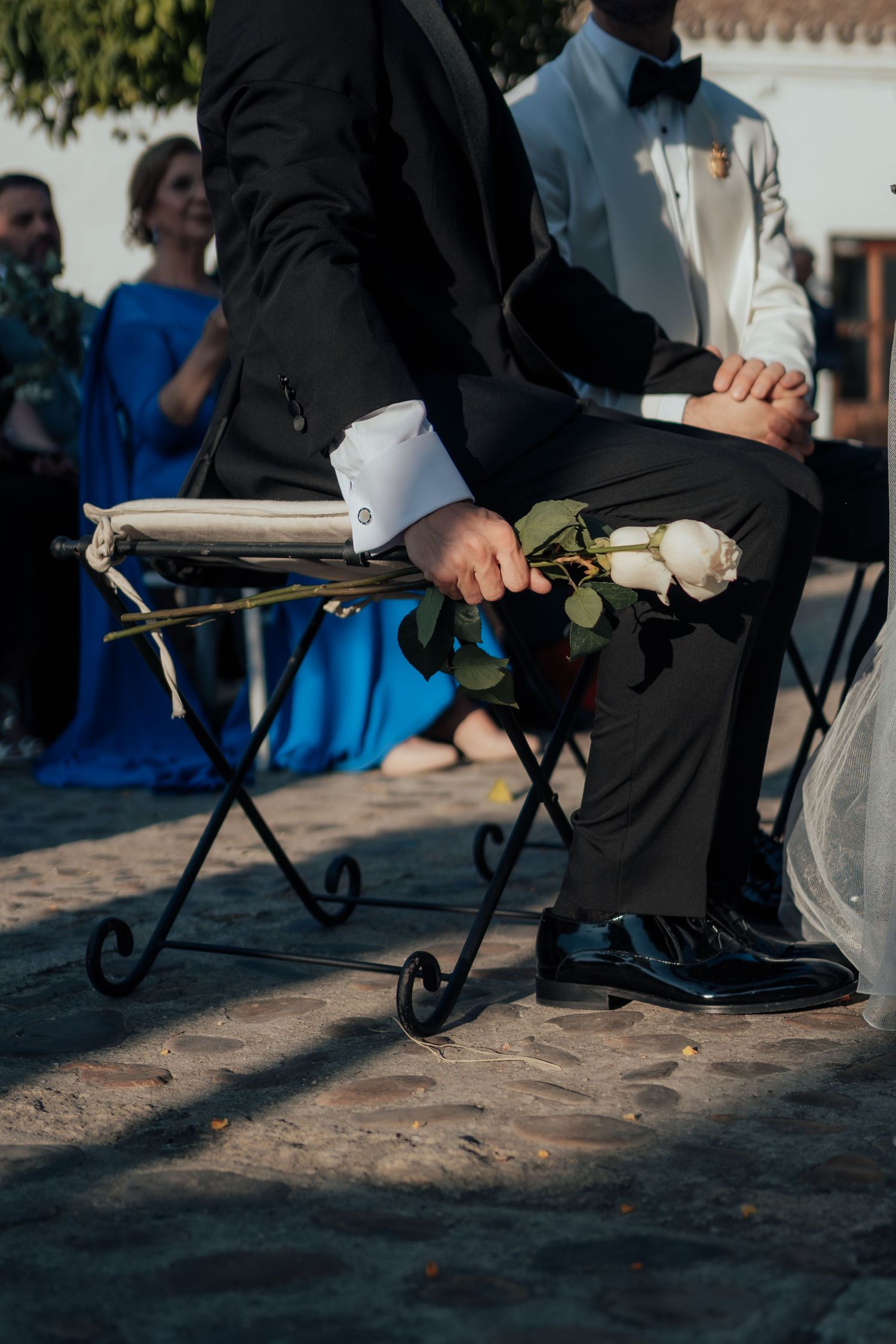 Groom in black tuxedo seated at outdoor wedding ceremony holding a single white rose.