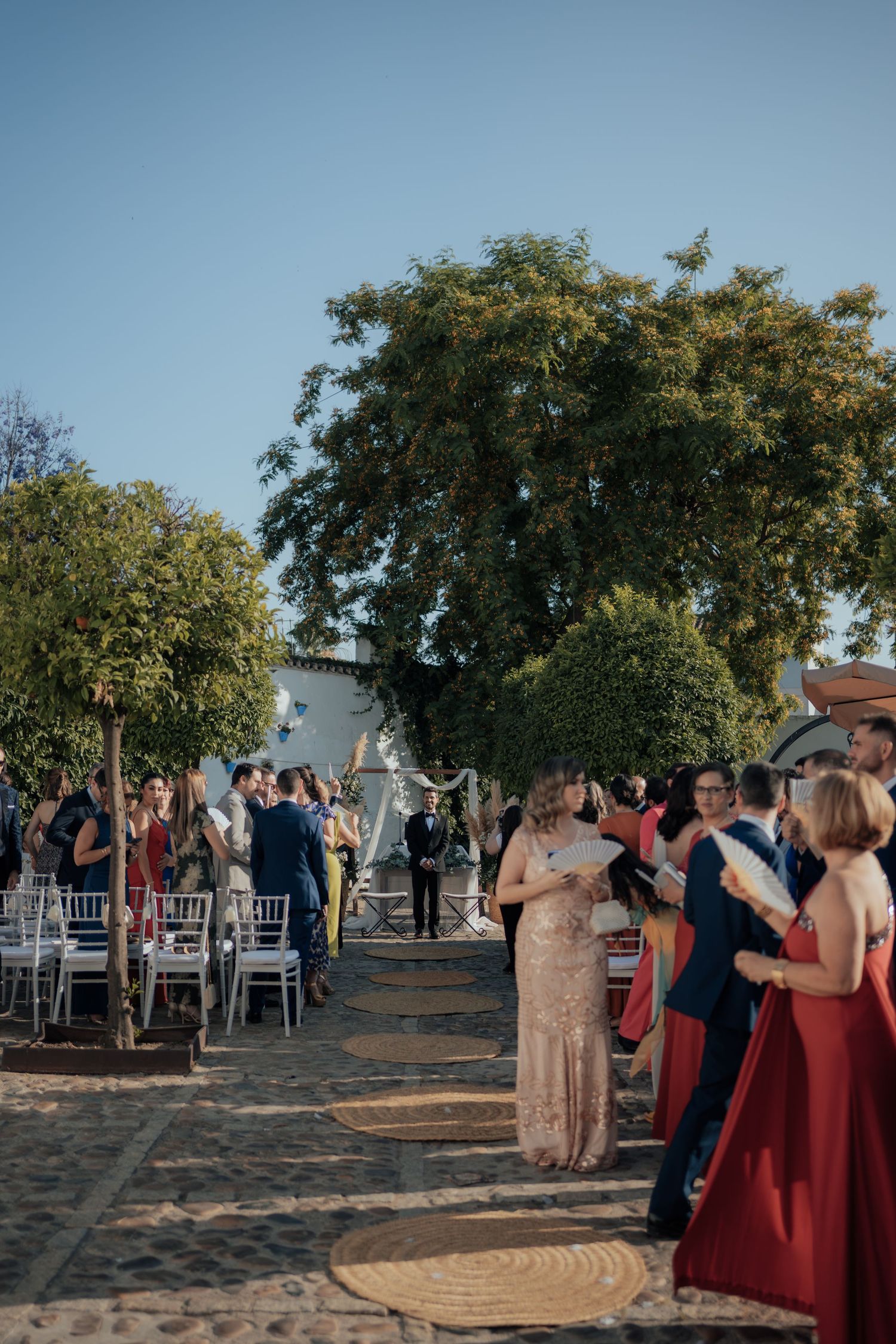 Wedding guests in formal attire line the aisle of an outdoor ceremony under clear blue skies.
