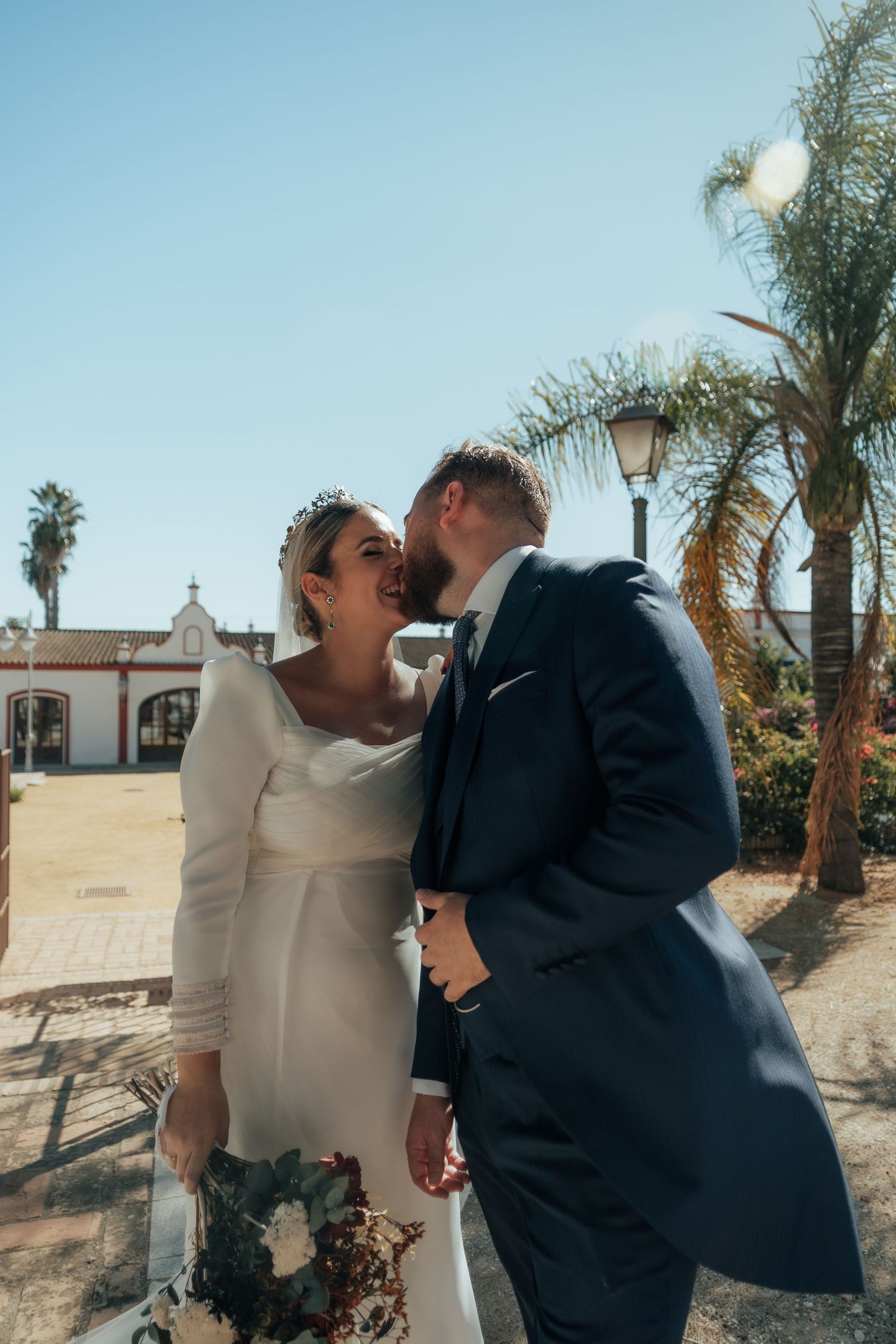 Groom in navy suit kisses smiling bride wearing a tiara and white gown under a sunny blue sky.